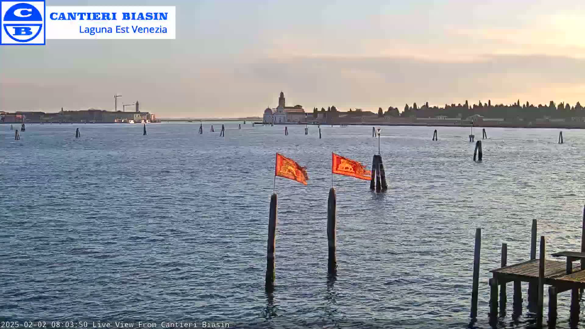 Two orange flags flutter on wooden posts in calm water, with a distant cityscape visible under a clear, pale sky.