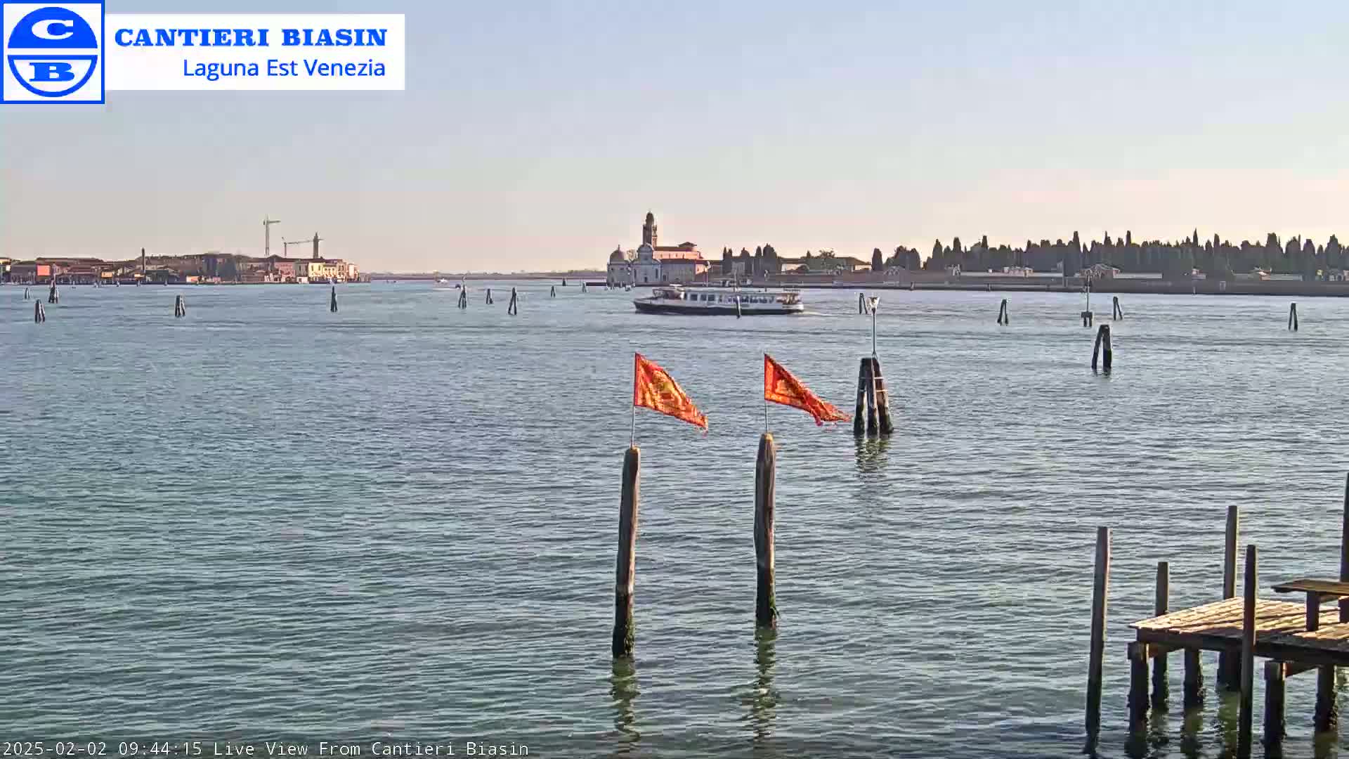 A calm body of water shows a distant building and a boat under a clear sky, with orange flags marking wooden posts in the foreground and a small dock to the lower right.