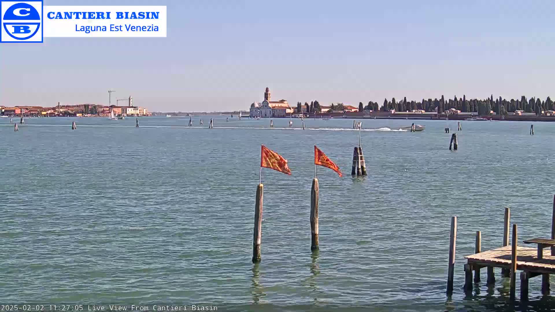A body of water shows a distant island with buildings and trees under a clear sky, two orange flags on wooden posts in the foreground, and a small boat on the water.