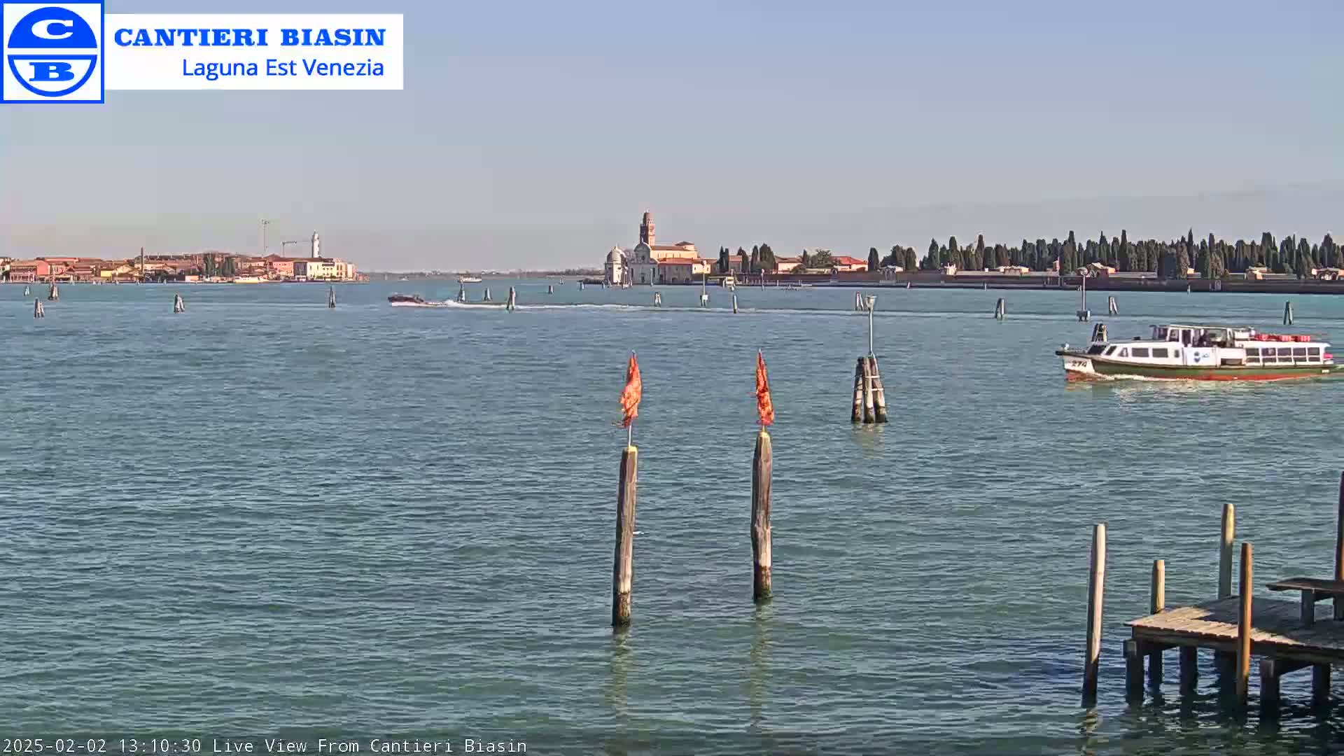 A calm, sunny day shows a waterway with a distant island featuring buildings and trees, a small passenger boat, and a wooden dock in the foreground.