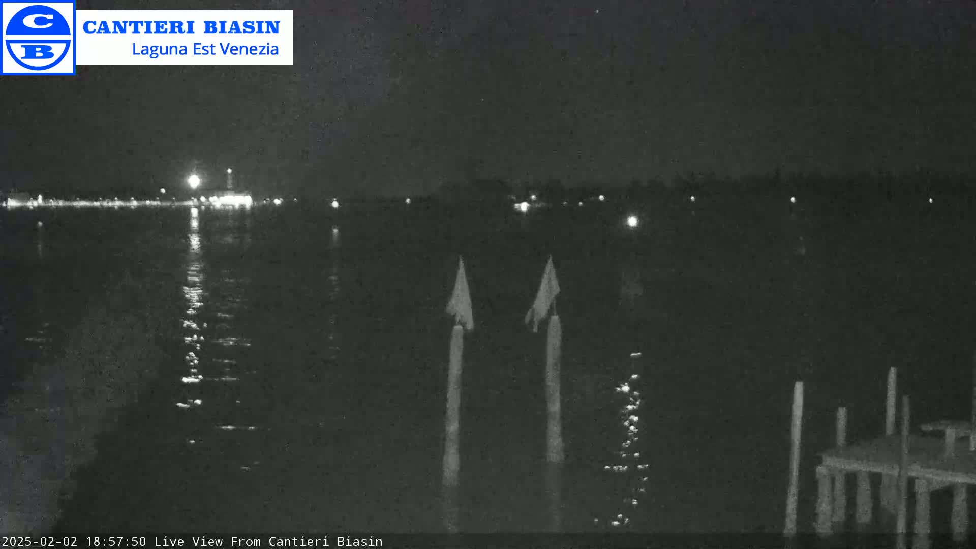 A nighttime view of a calm body of water with lights visible in the distance and two posts in the foreground.