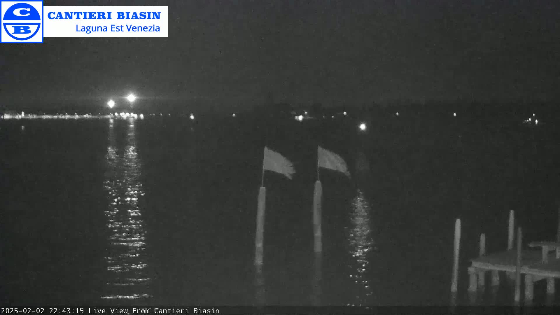 A nighttime view of calm water reflecting distant lights, with two flags on poles near a small dock.