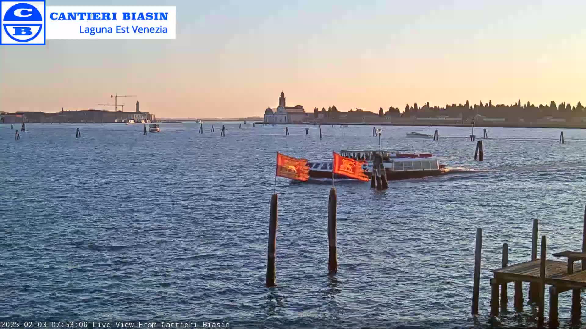 A calm body of water at sunset shows a long boat with orange flags and several other small boats, with a distant island and buildings in the background.