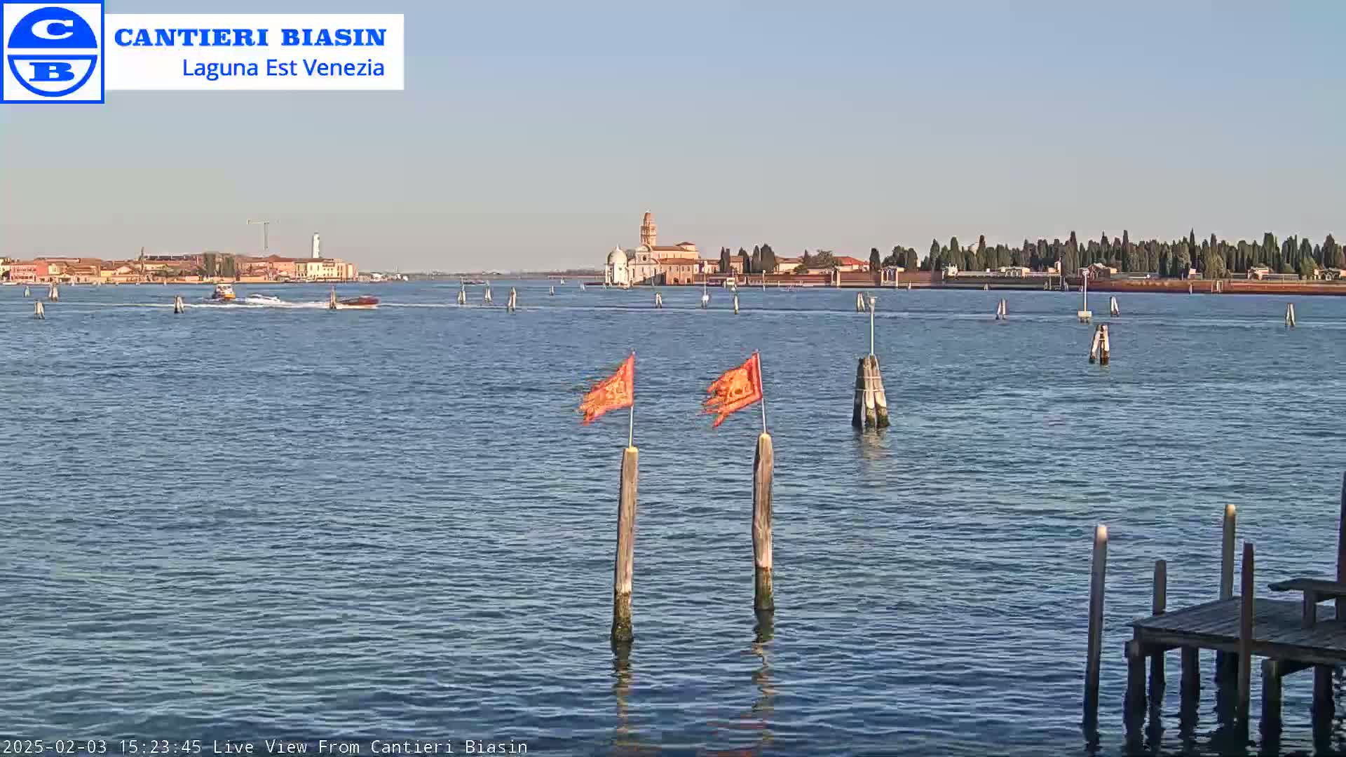 A calm body of water shows several boats, a distant island with buildings, and wooden posts with orange flags in the foreground under a clear sky.