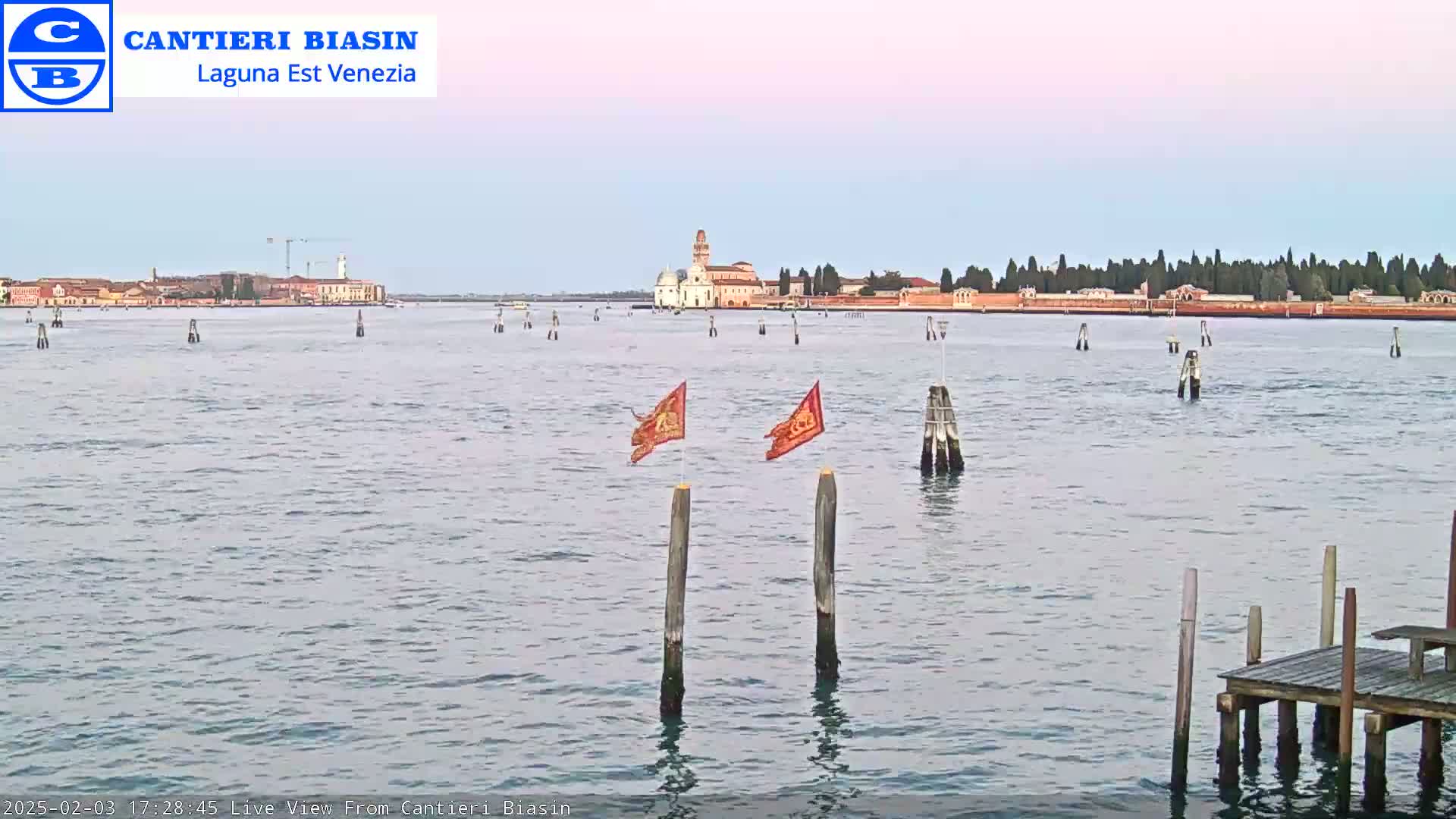 A calm body of water shows a distant island with buildings under a pale pink sky, with two small flags on posts in the foreground and a wooden dock to the lower right.