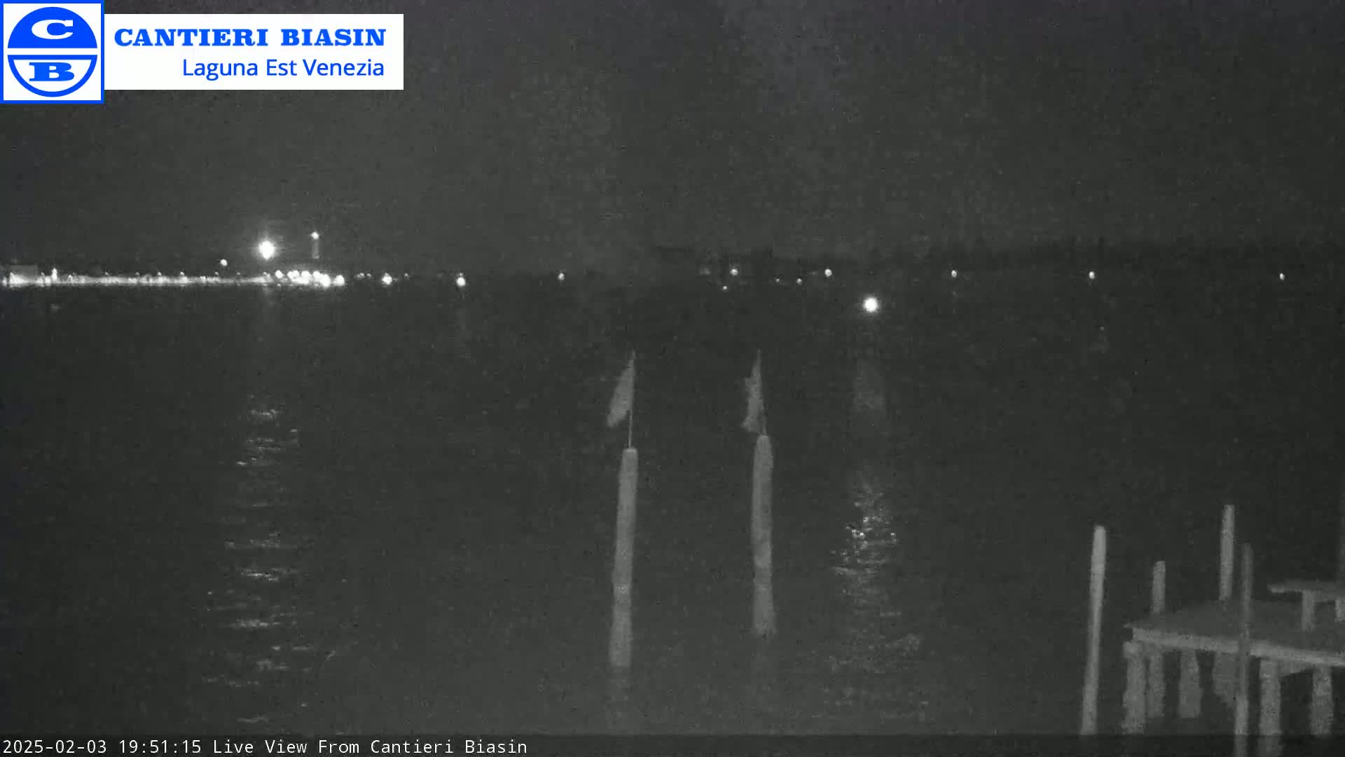 A nighttime view of a dark body of water with dimly lit structures on the far shore and several posts in the foreground under a clear sky.