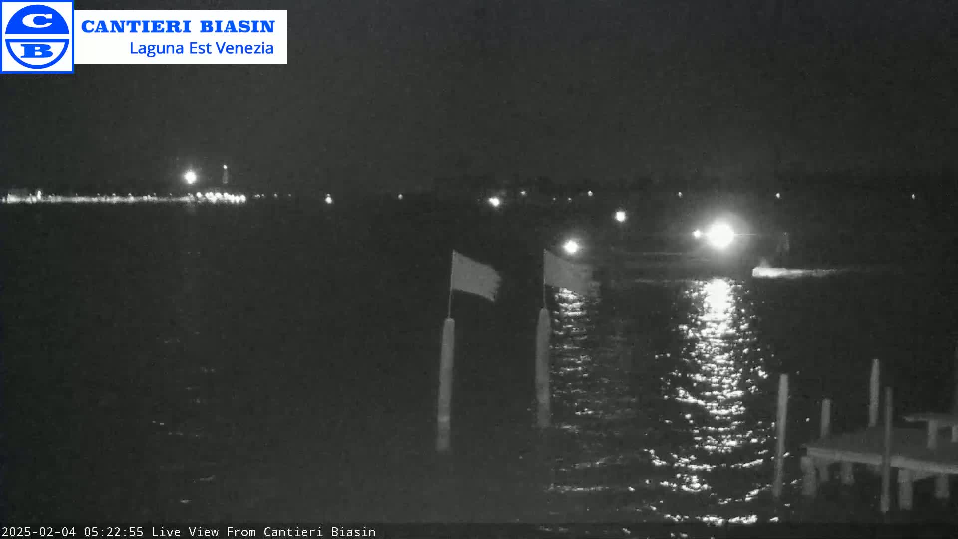 At night, a dark body of water reflects lights from a distant shore and a vessel, while two flags are visible on posts in the foreground.