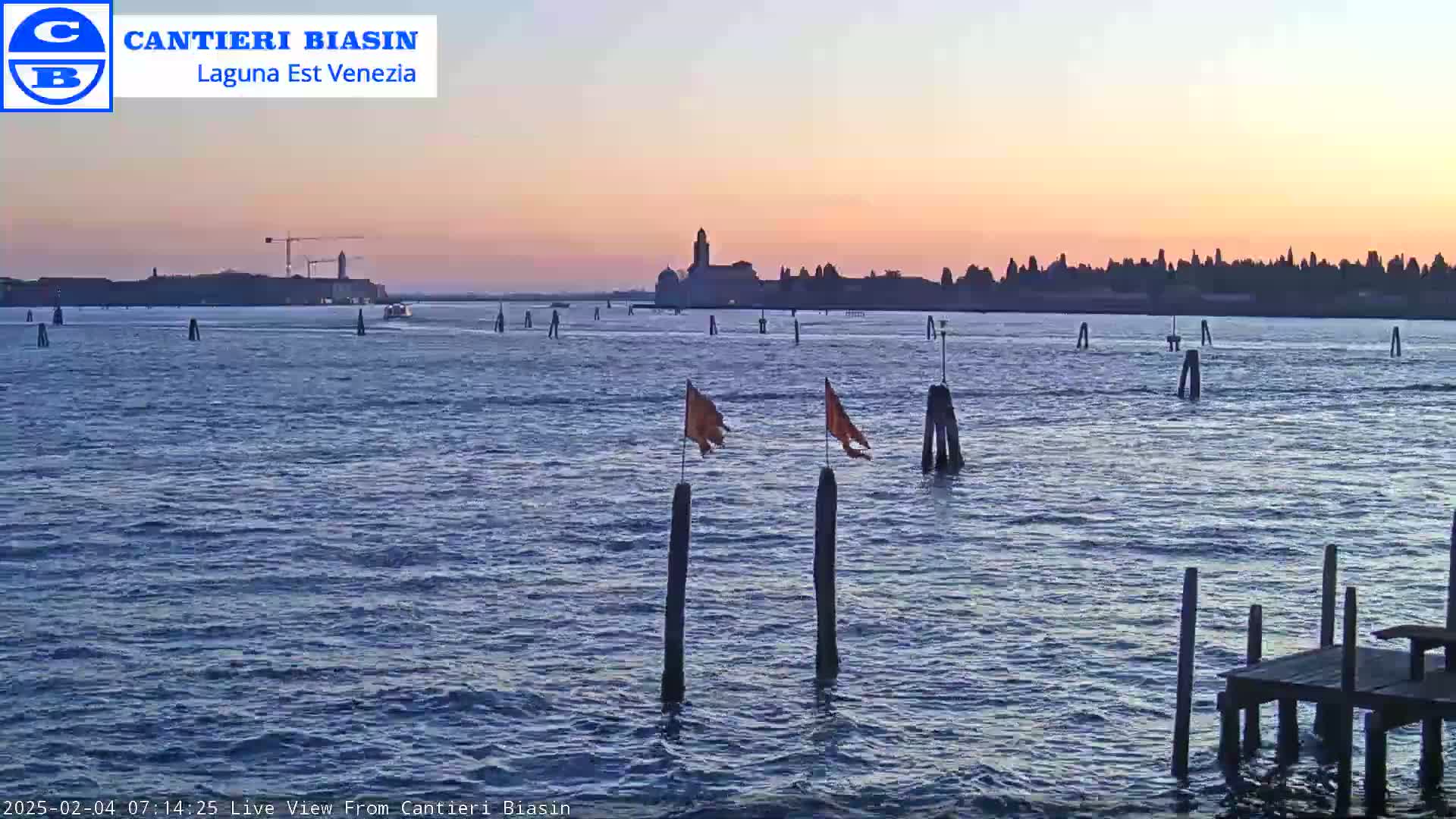 A calm body of water at sunset shows a distant cityscape silhouetted against a pale orange and pink sky, with a few boats and wooden posts in the foreground.