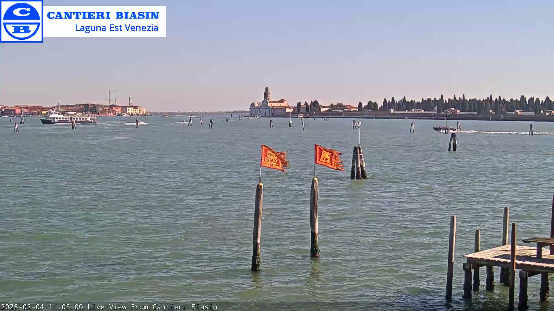 A calm, sunny day shows a waterway with boats, a distant island with buildings, and wooden posts with orange flags in the foreground.