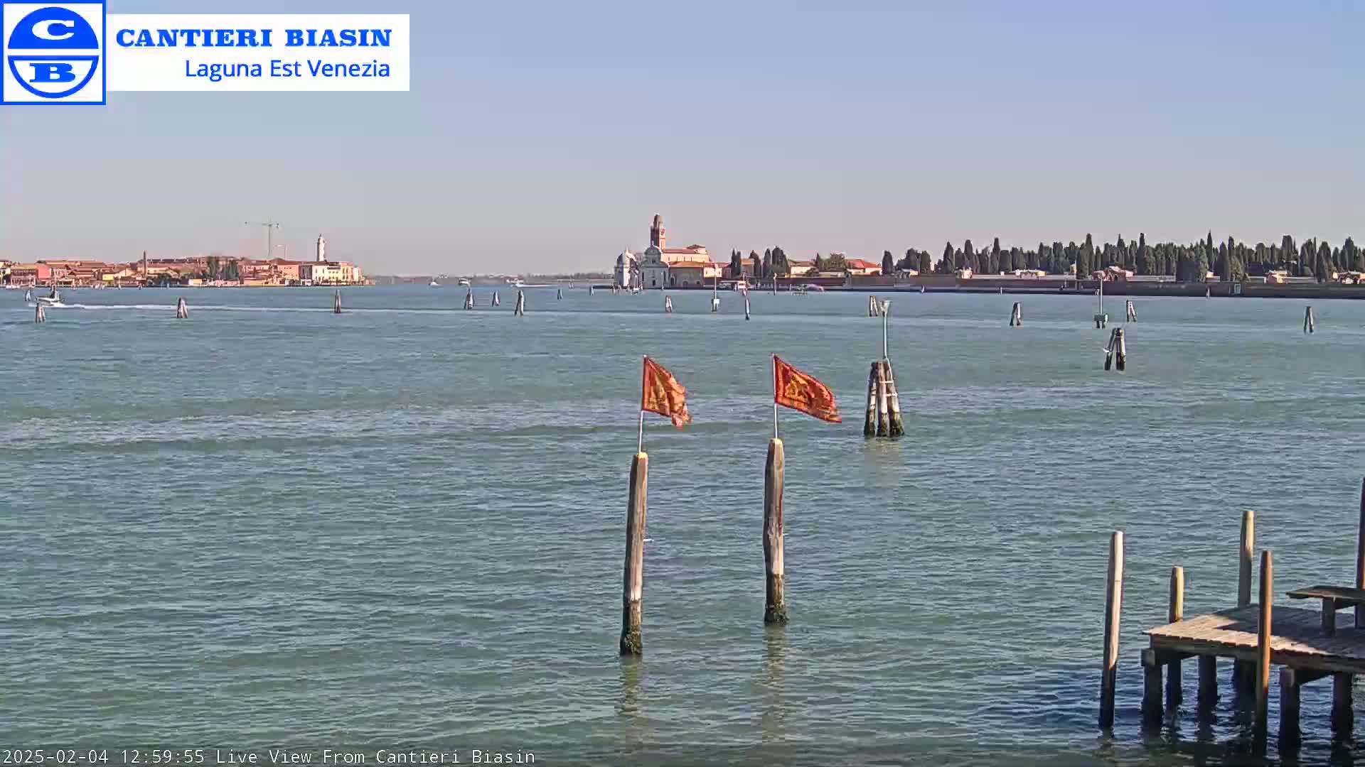 A calm body of water shows a distant island with buildings and trees under a clear sky, with two orange flags on wooden posts in the foreground near a small dock.