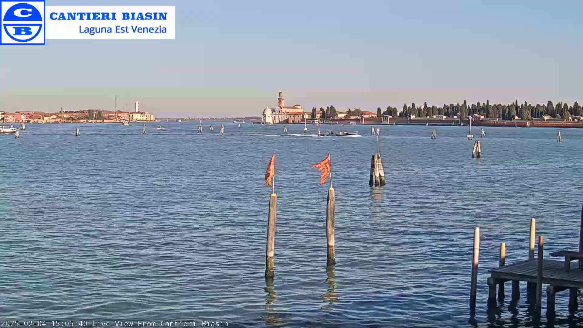 A calm body of water shows a distant shoreline with buildings under a clear sky, featuring several wooden posts with orange flags in the foreground.