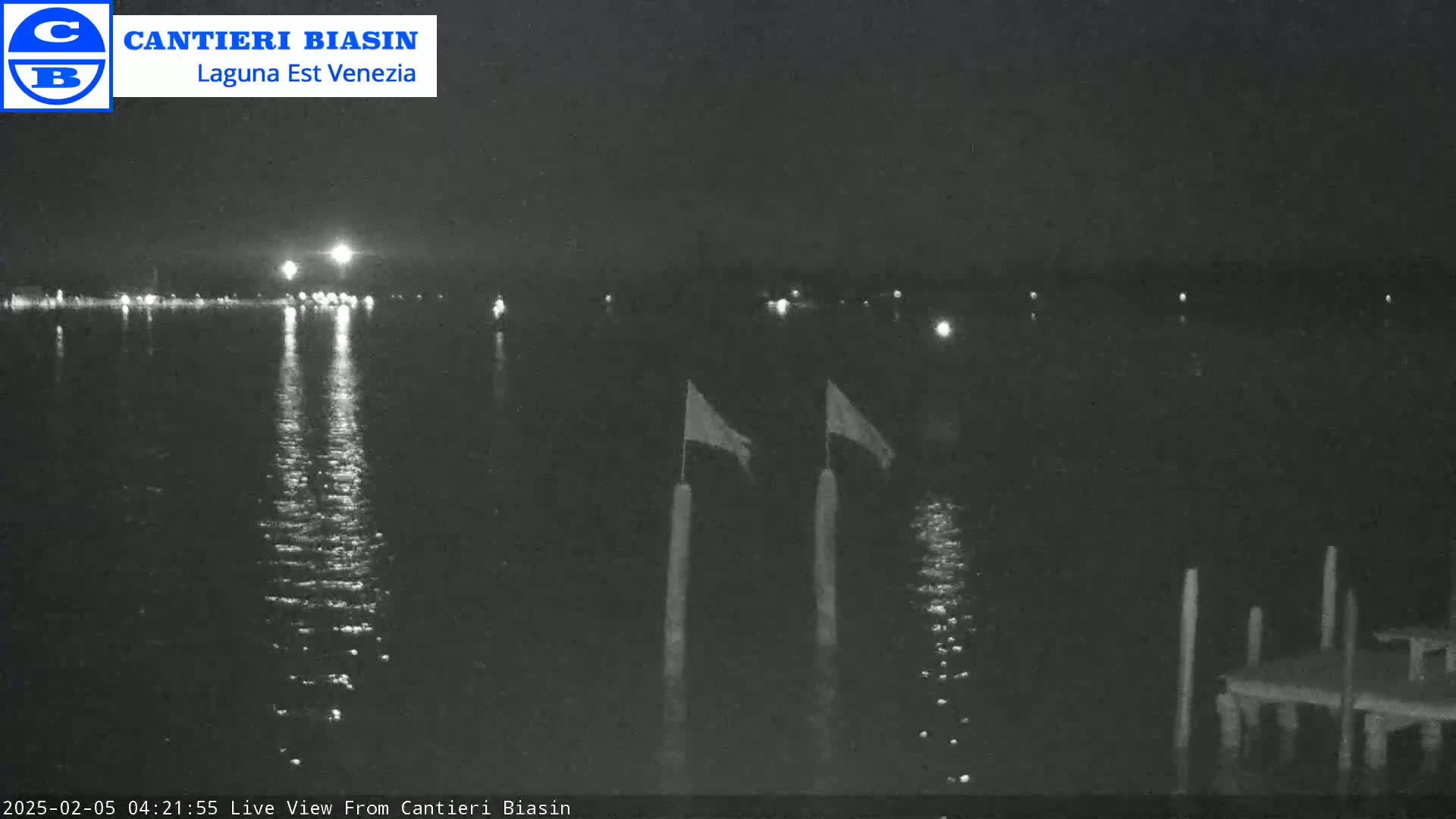 A nighttime view of calm water reflecting lights from a distant shoreline, with two flags on poles in the foreground and a small dock to the right.