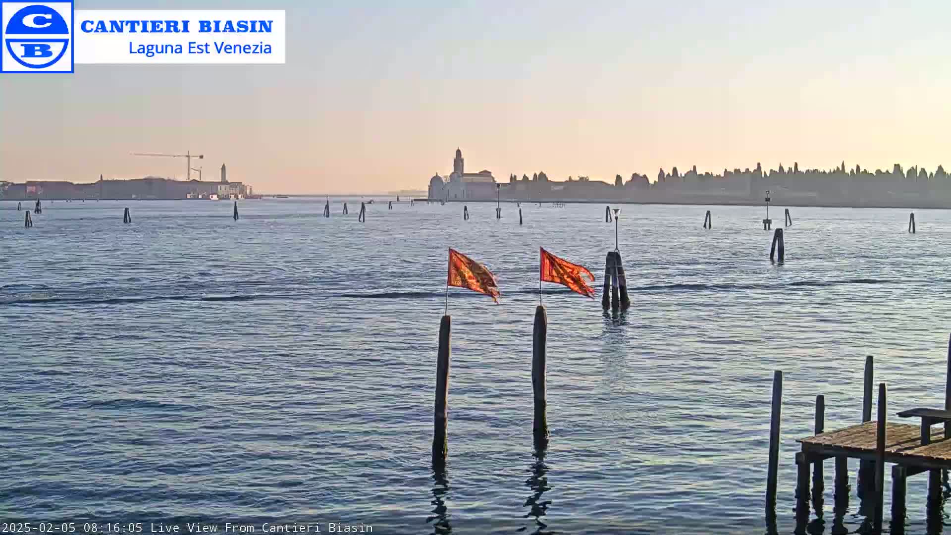 A calm body of water with several posts and orange flags, a distant city skyline, and a small wooden dock under a clear, light-colored sky.