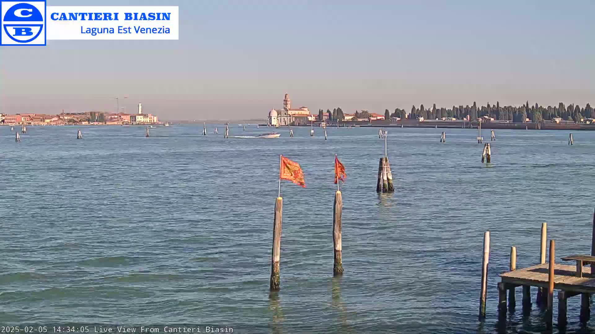 A calm body of water with several wooden posts and small orange flags, a distant island with buildings, and a small boat in the distance under a clear sky.