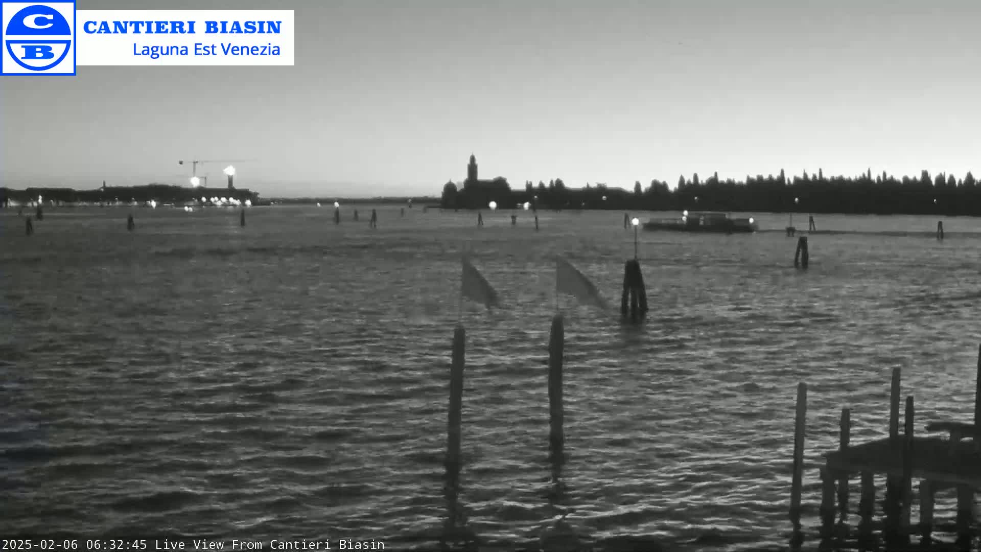 A grayscale image shows a calm body of water with a distant shoreline, several poles in the water, and a small boat in the mid-ground, all under a twilight sky.