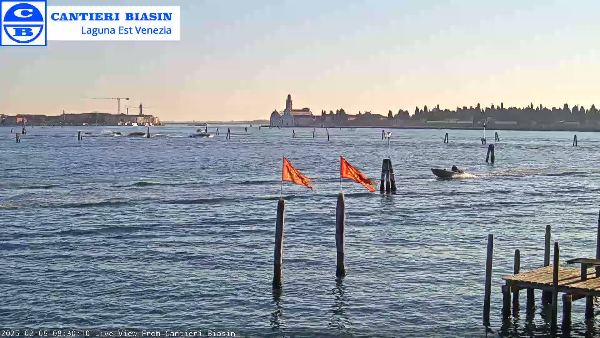 A calm body of water with a small motorboat and orange flags on posts,  a distant island with buildings, and a clear, sunny sky.