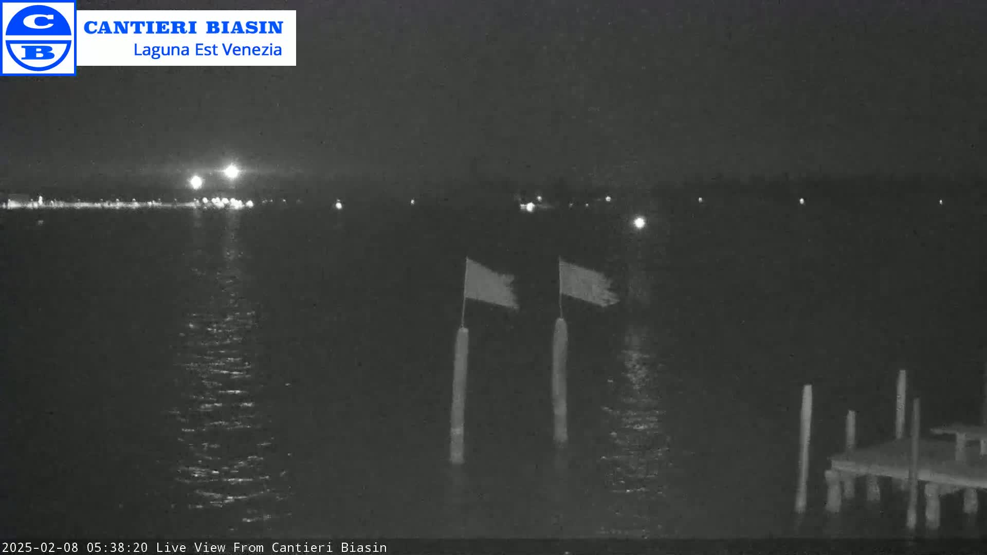 A nighttime view of calm water shows a distant shoreline with lights, two flags on poles in the foreground, and a small dock to the right.