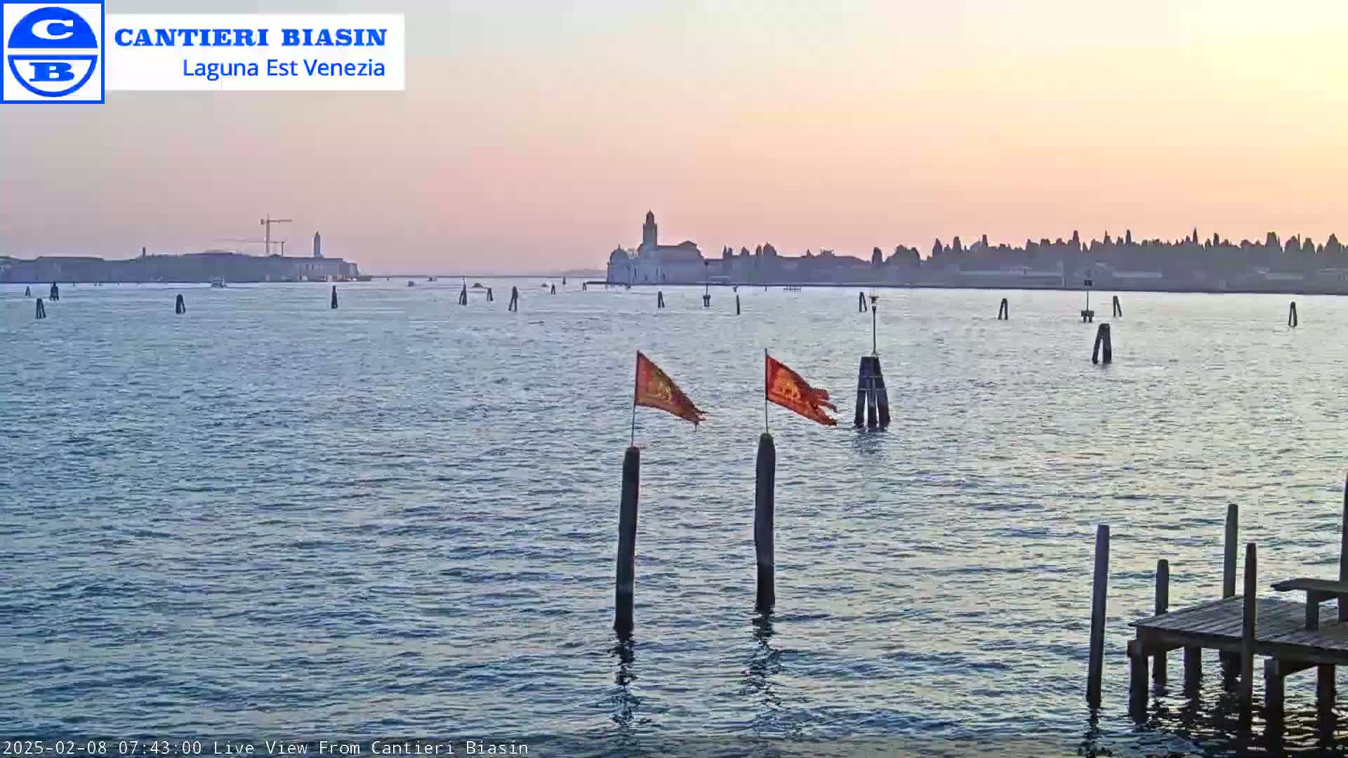 A calm body of water at sunset, showing a distant cityscape and two orange flags on posts in the foreground.