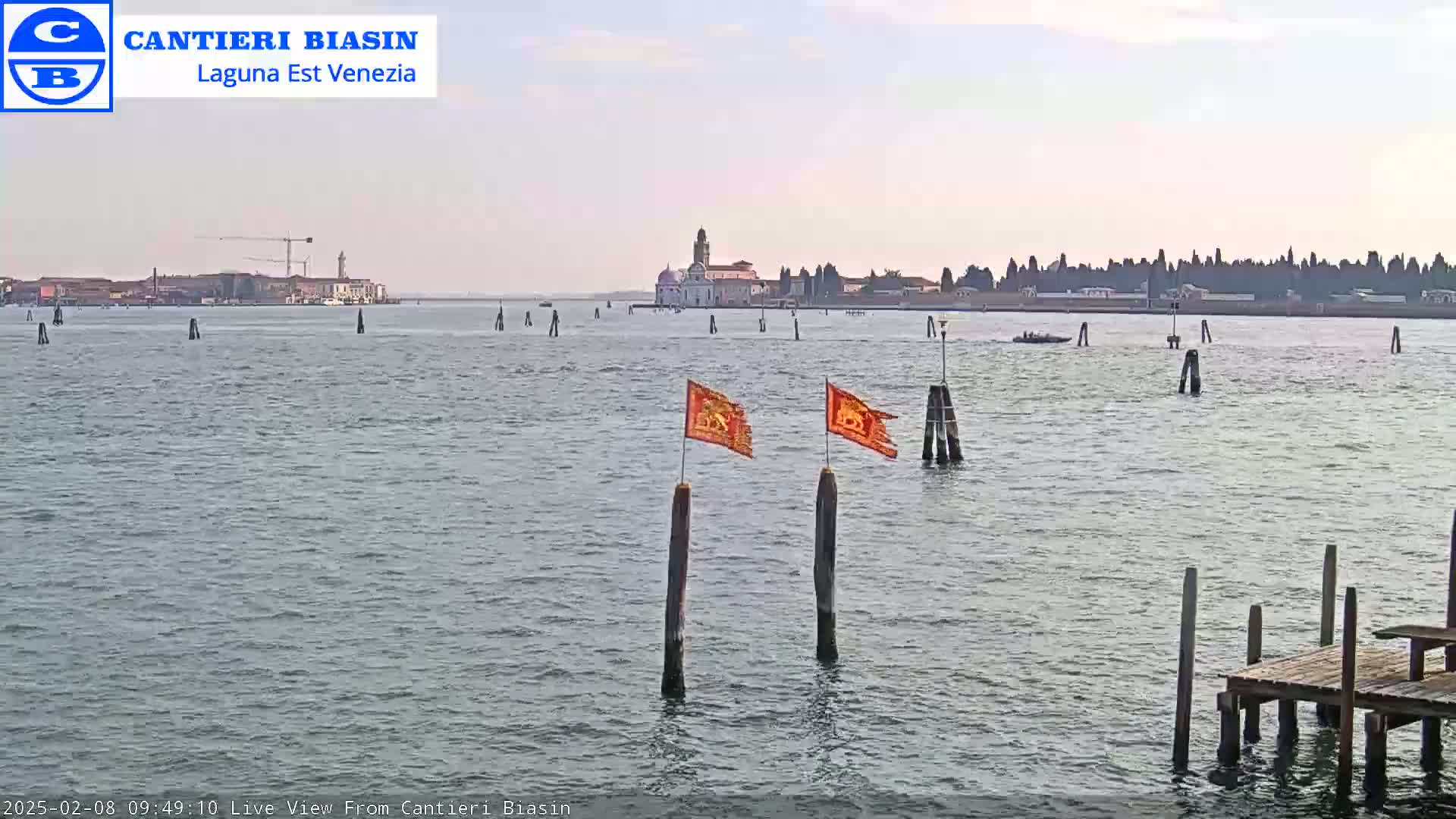 A calm body of water shows a distant cityscape and island with a church, two orange flags on posts in the foreground, and a wooden dock to the lower right.
