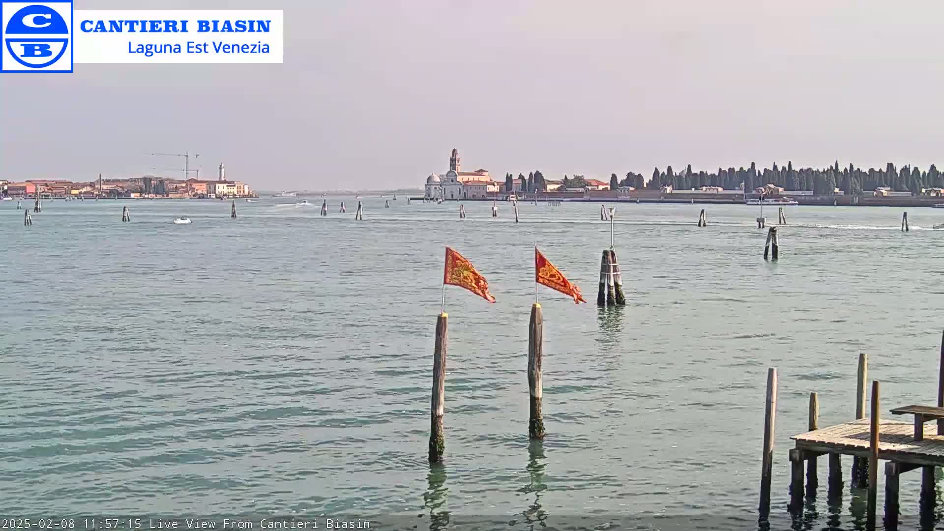 A calm body of water shows a distant island with buildings, a few boats, and wooden posts with orange flags in the foreground under a hazy sky.