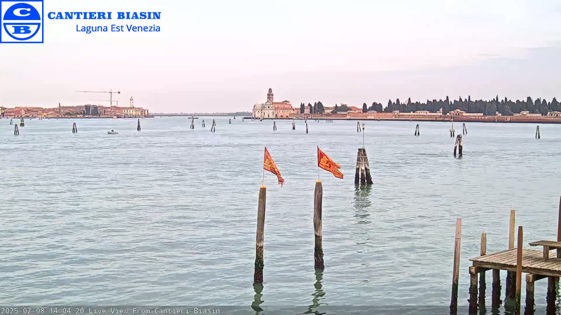 A calm body of water shows a distant island with buildings and trees under an overcast sky, with two orange flags on wooden posts in the foreground and a small dock to the lower right.