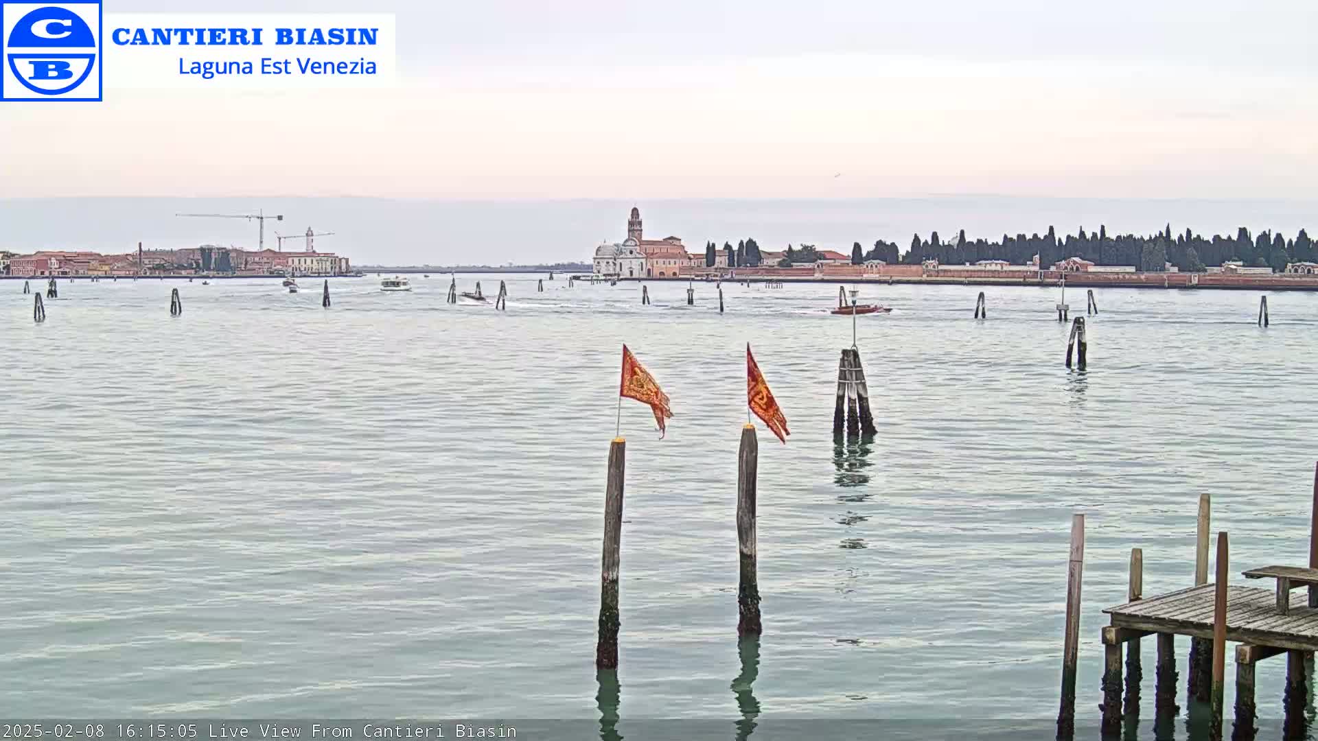 A calm body of water shows a distant island with buildings, a few boats, and two orange flags on wooden posts in the foreground under an overcast sky.