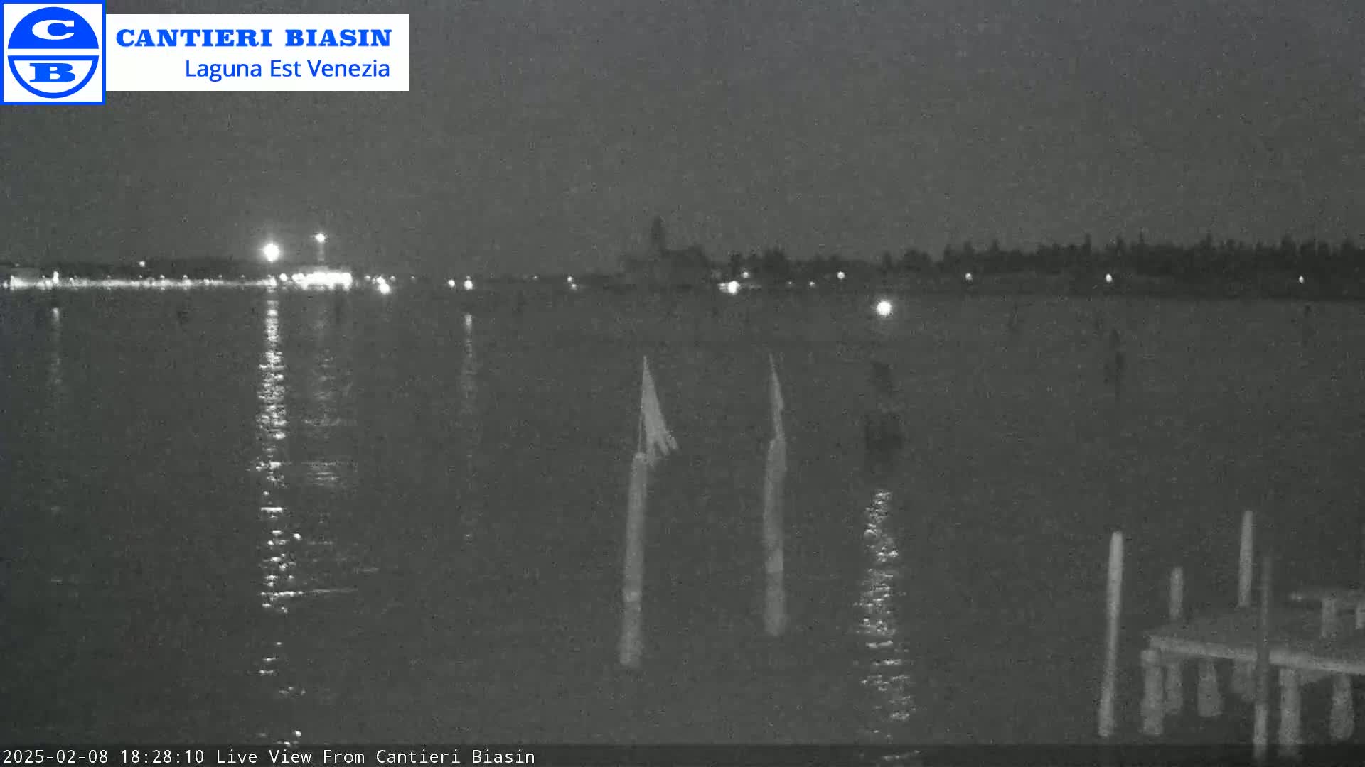 A nighttime view of calm water with a distant shoreline showing lights, and a few posts in the foreground.