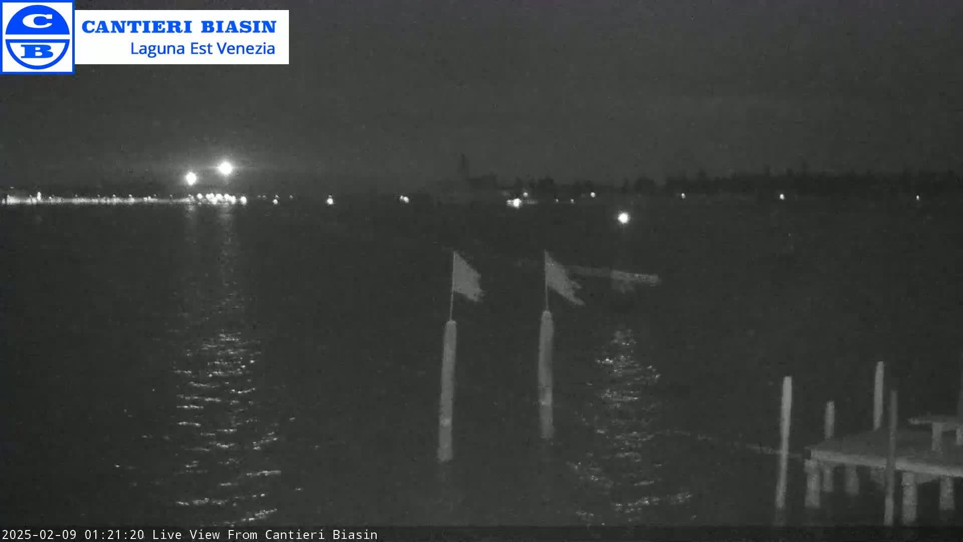 A nighttime view of calm water with a distant shoreline showing faint lights, and a small dock with flags in the foreground.