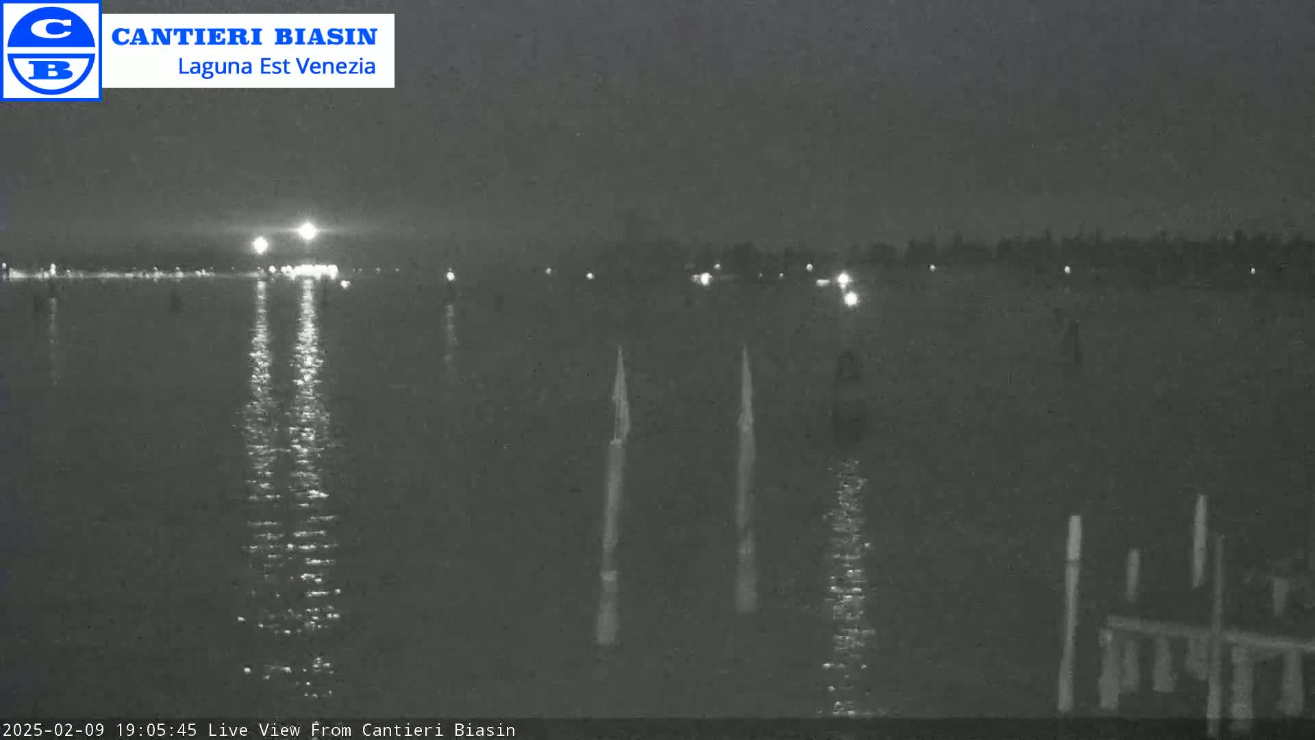 A nighttime, low-light view of calm water reflecting lights from a distant shore, with several vertical posts visible in the water near the foreground.
