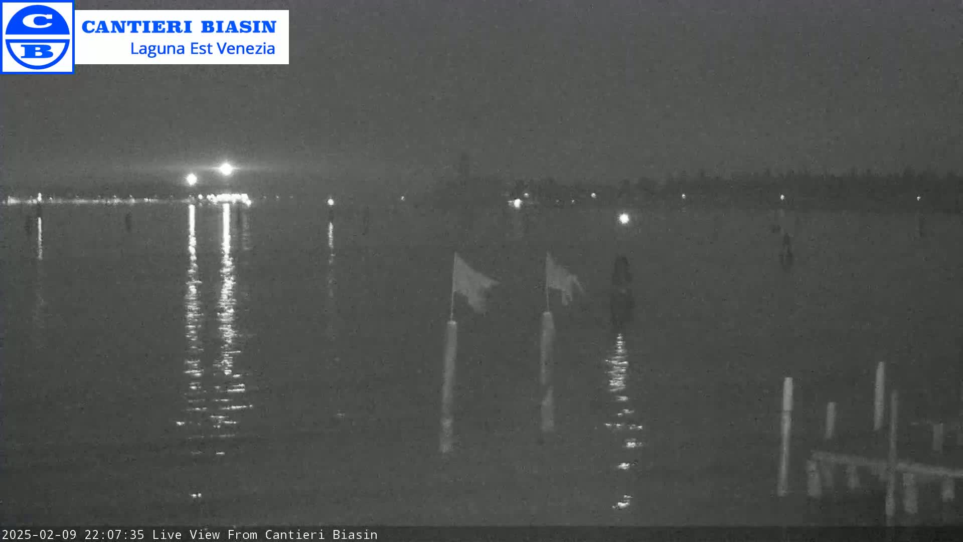 A nighttime view of a calm body of water with lights visible on the opposite shore and two flags on posts in the water.