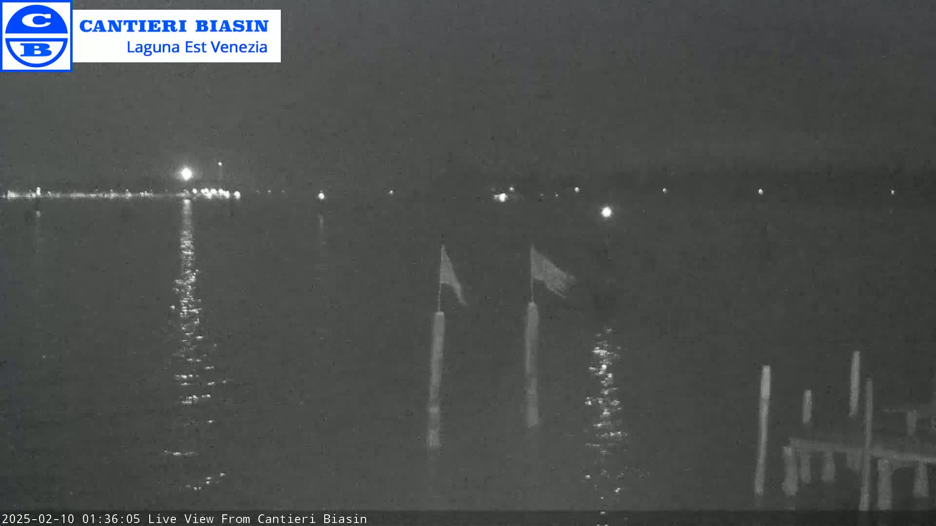 A nighttime view of calm water with a few lights on the far shore and flags on posts in the foreground.