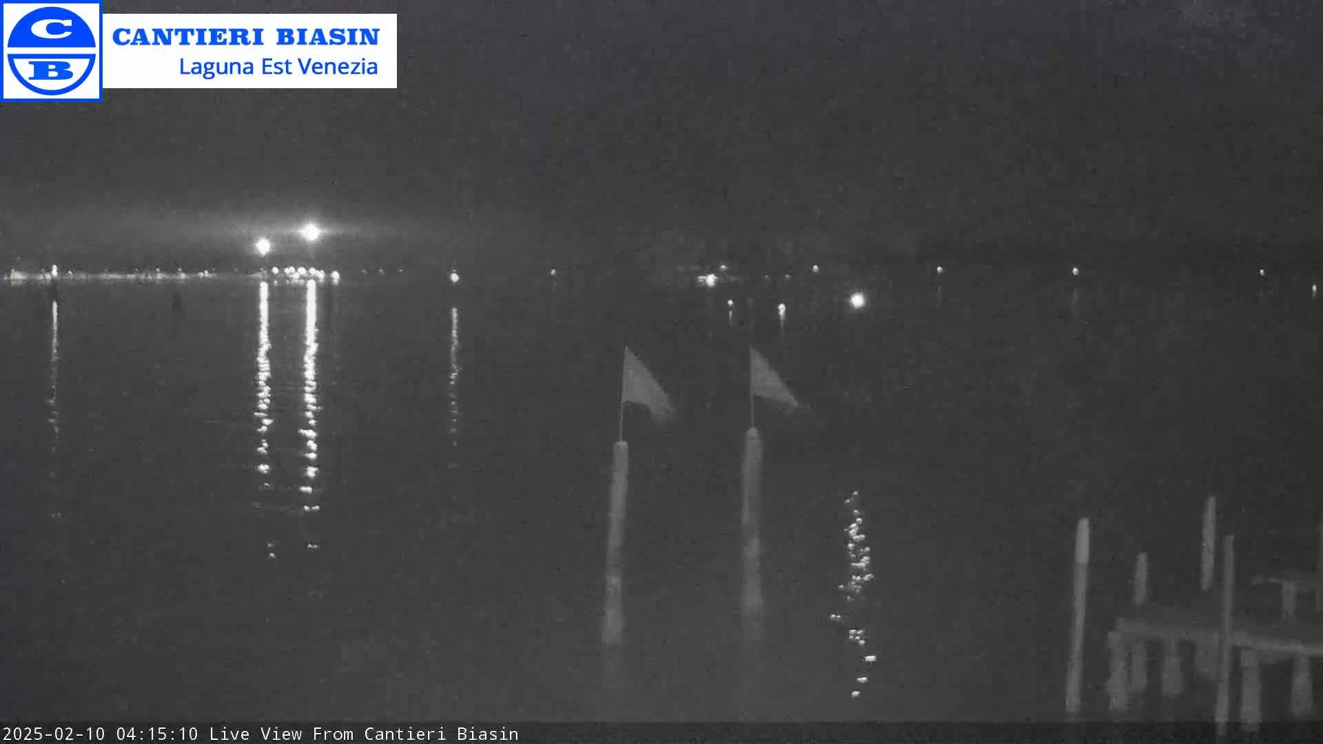 A nighttime view of calm water reflecting distant lights, with two flags on poles in the foreground.
