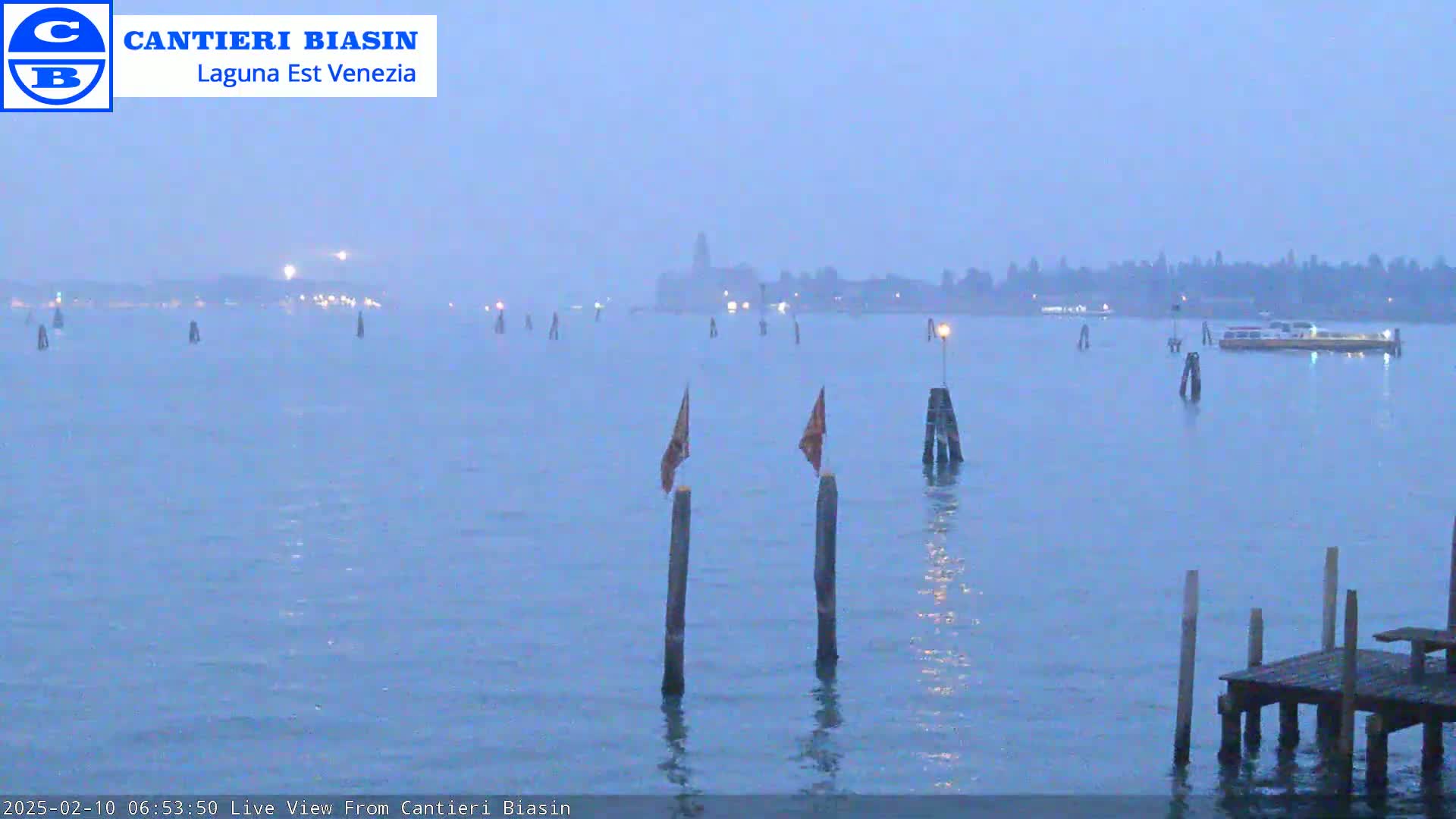 A hazy, light-blue body of water shows a distant shoreline and a few boats, with several wooden posts and a small dock in the foreground.