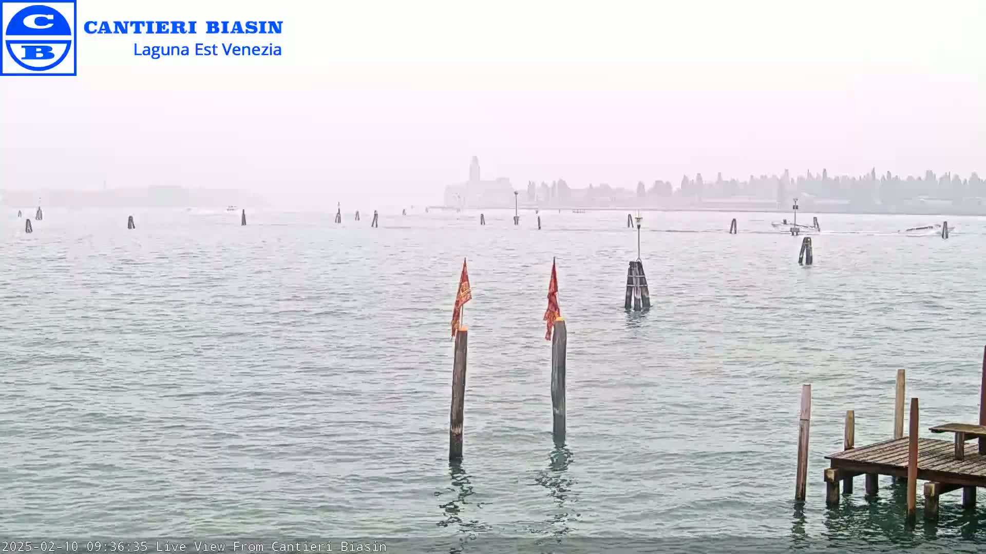 A hazy, calm body of water with several posts and markers, a distant city skyline, and a small wooden dock in the foreground.