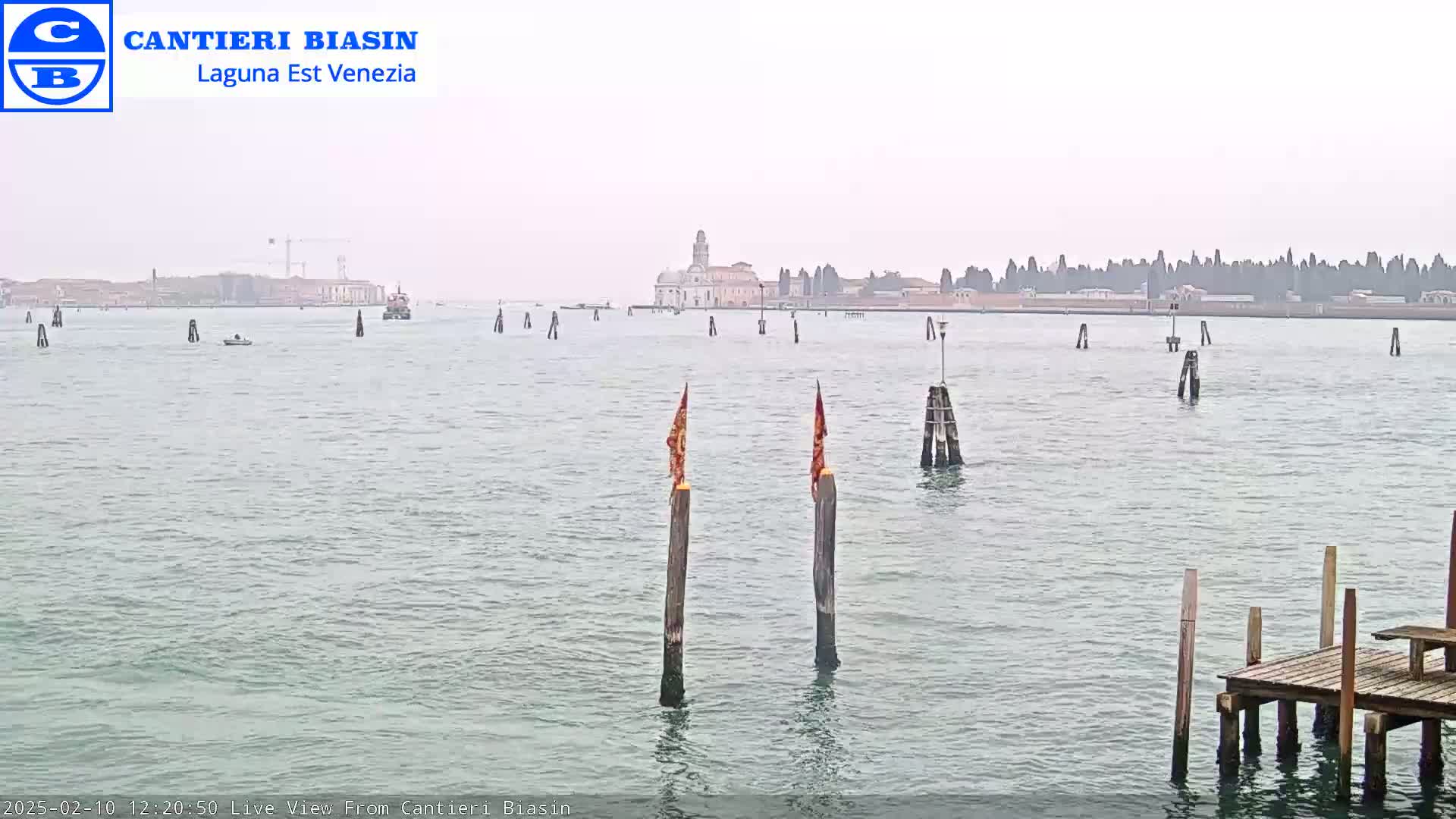 A hazy view of a calm body of water shows a distant island with buildings and trees, wooden posts in the foreground, and a small wooden dock to the lower right.
