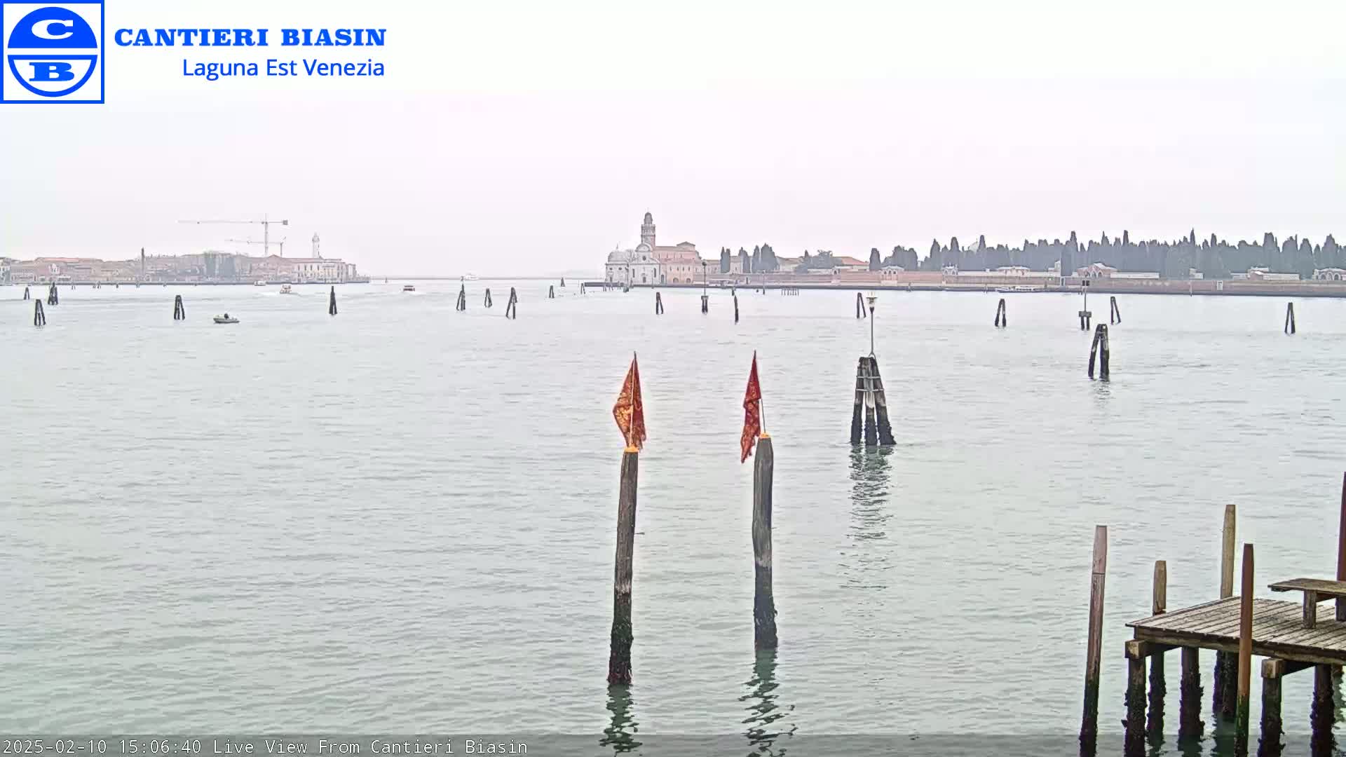A calm body of water with wooden posts and small flags, showing a distant cityscape under an overcast sky.