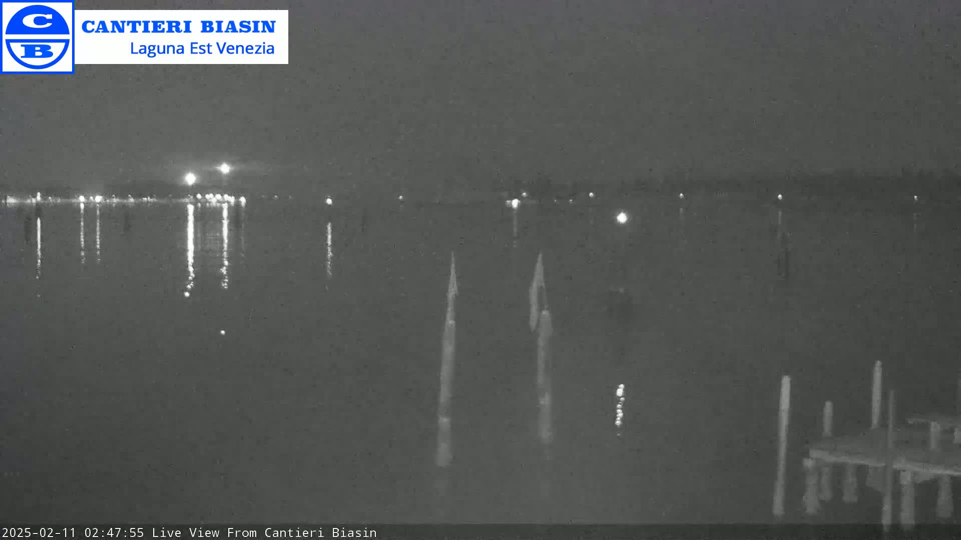 A nighttime view of calm water reflecting distant lights, with several posts and a dock visible in the foreground under a dark sky.