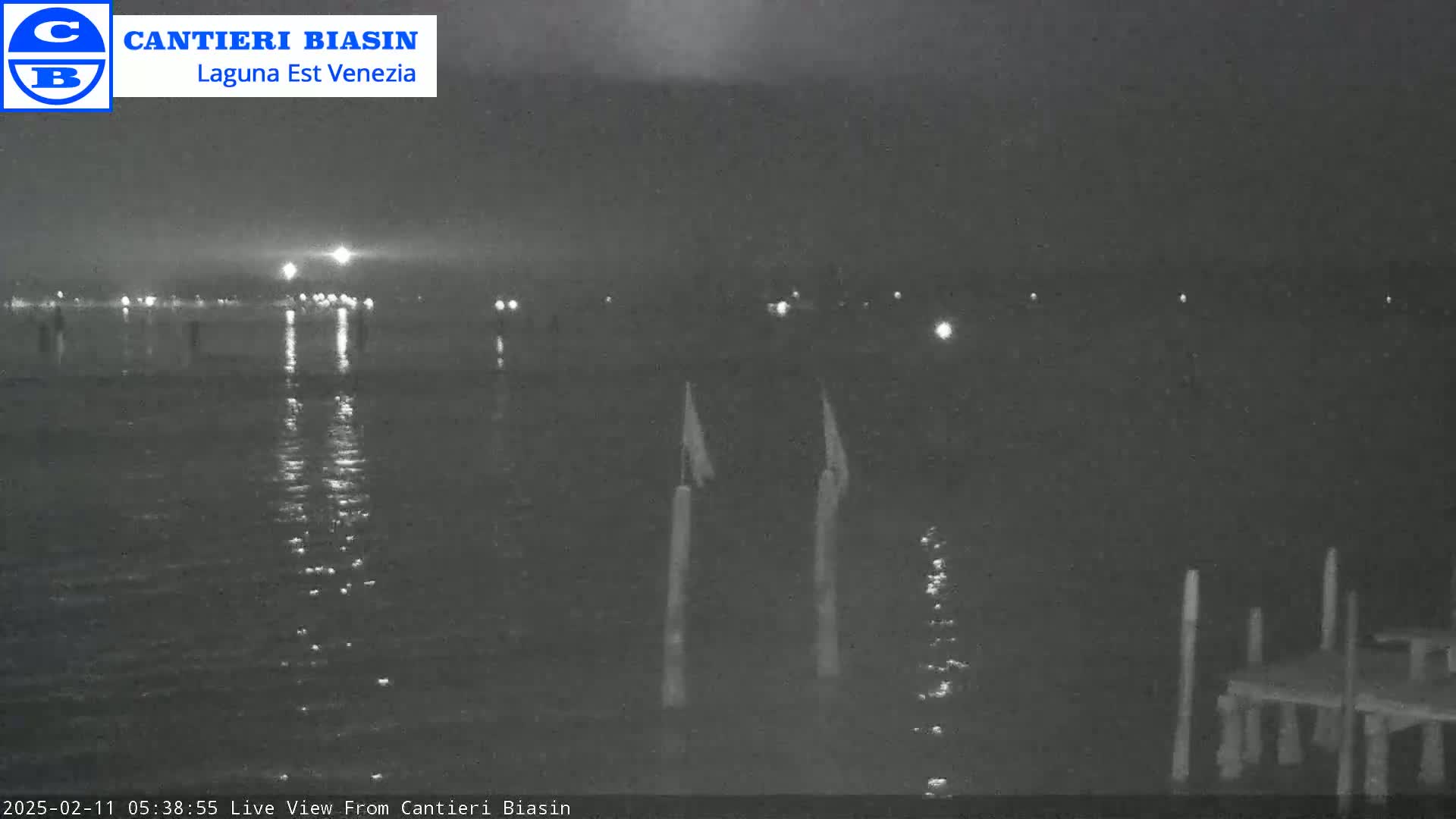 A nighttime view of calm water with distant lights reflecting on the surface, and several posts in the foreground.