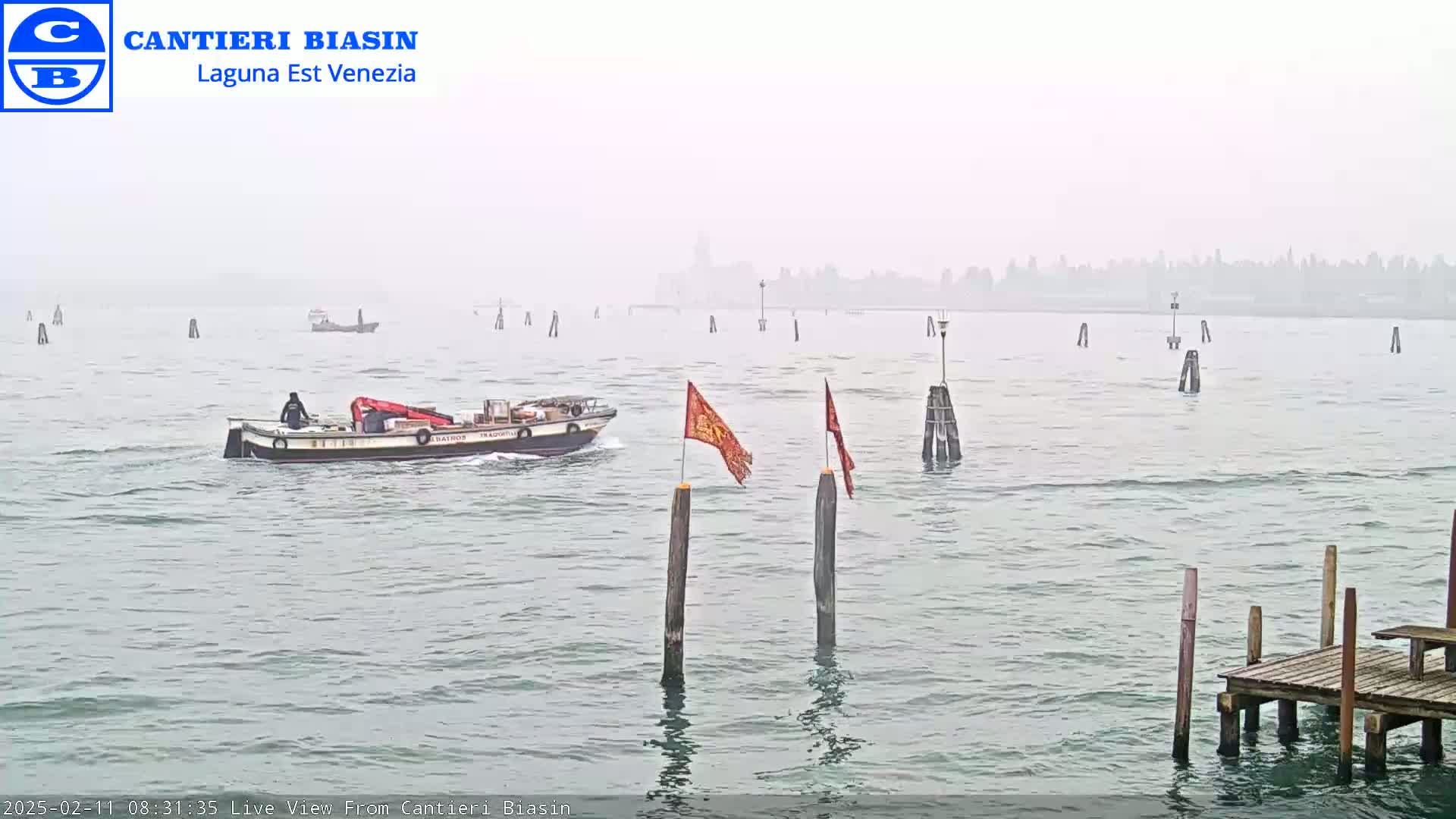 A boat travels on a foggy waterway past wooden pilings and flags, with a distant hazy city in the background.