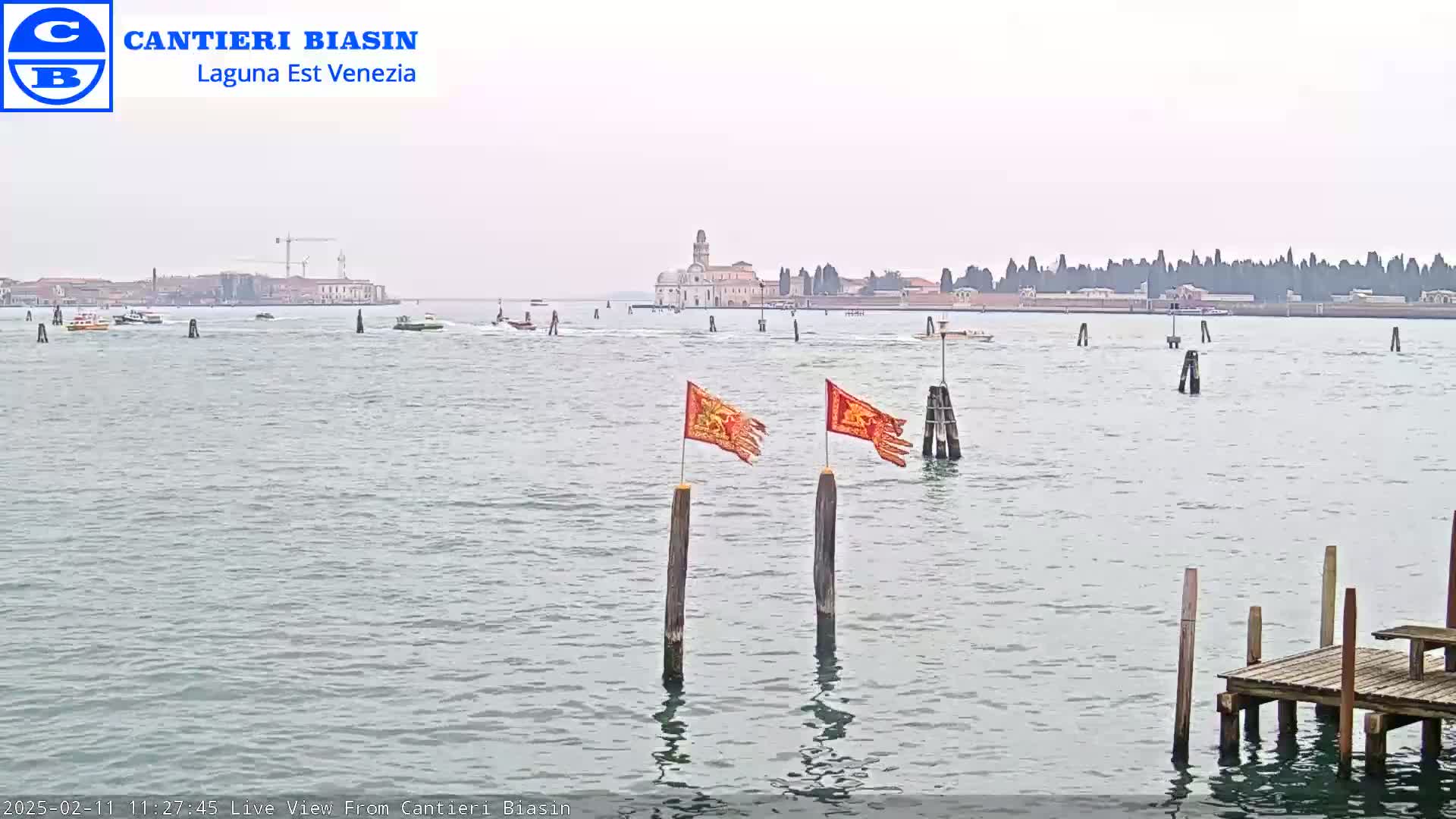 A calm body of water shows a distant cityscape under an overcast sky, with two orange flags on wooden posts in the foreground and a small dock to the lower right.