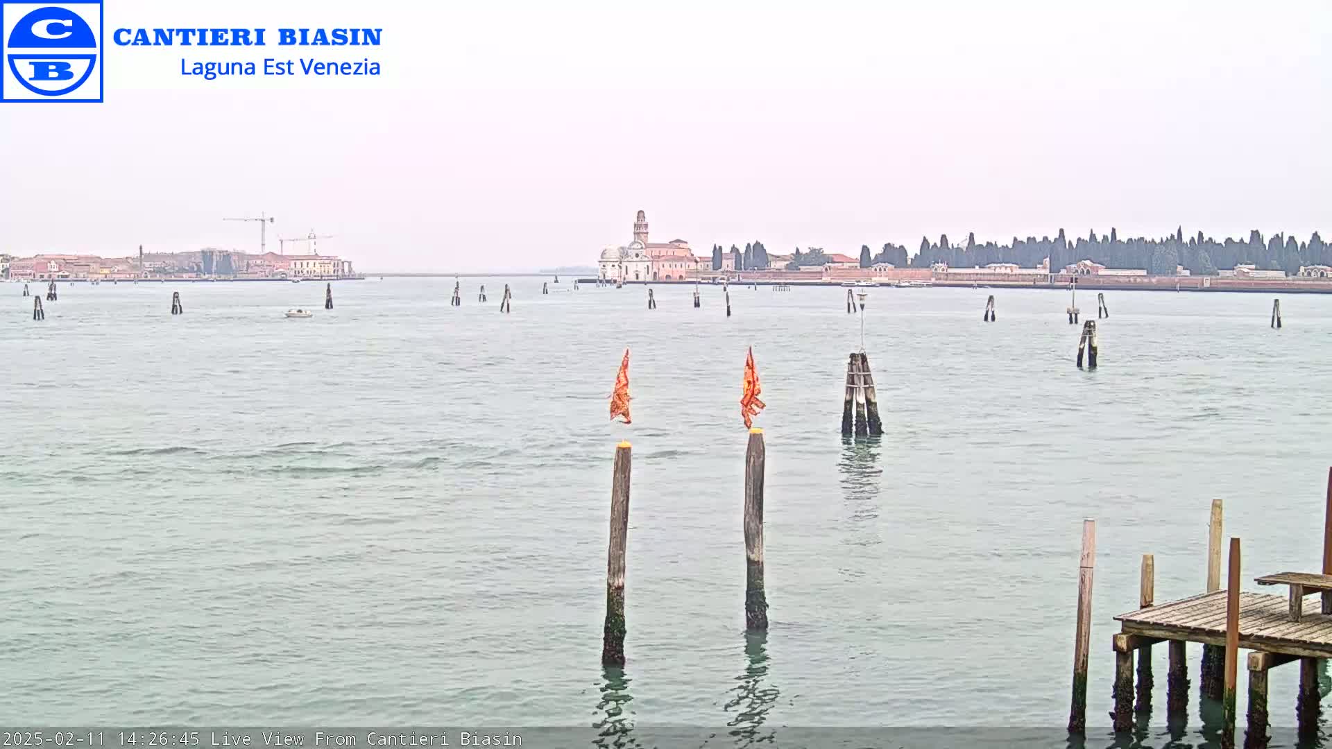 A calm body of water shows a distant island with buildings under an overcast sky, featuring several wooden posts and a small dock in the foreground.