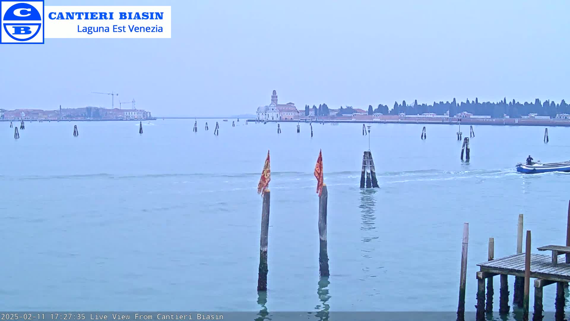 A calm, light-blue body of water shows a distant cityscape, several wooden posts with flags, a small boat, and a wooden dock in the foreground.