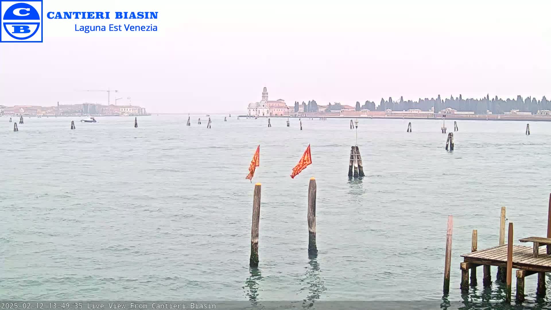 A hazy view across calm water shows a distant island with buildings and trees, a small boat, and wooden posts with orange flags in the foreground.