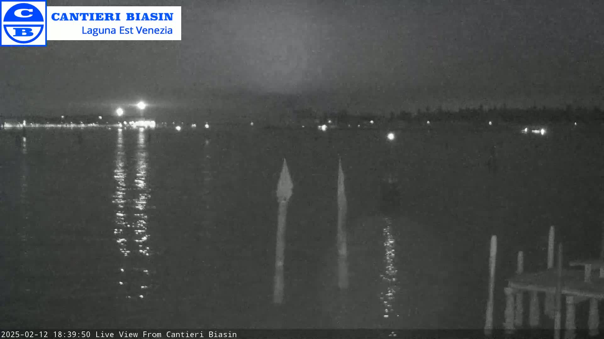 A nighttime view of calm water reflecting distant lights, with two tall, thin objects standing in the water in the foreground.