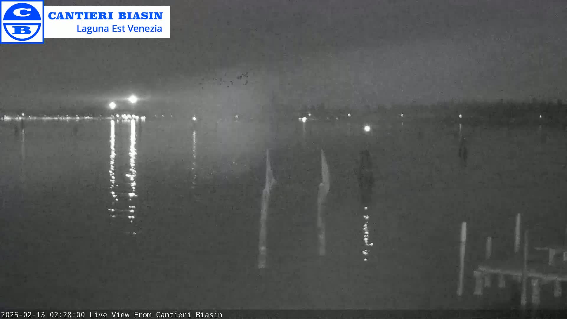 A nighttime view of calm water reflecting lights from a distant shore, with several posts visible in the foreground under a dark sky.
