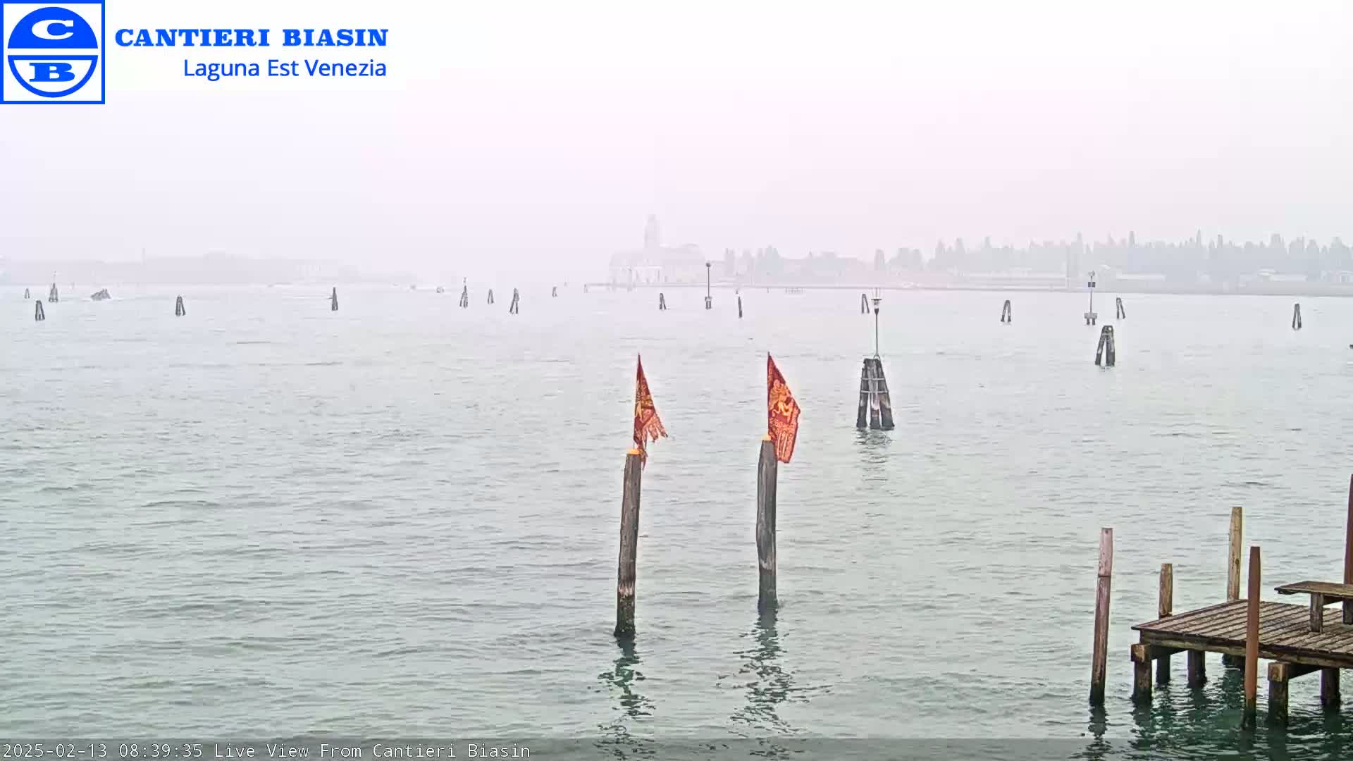 A foggy view of a calm body of water with wooden posts and flags, and a distant hazy cityscape.
