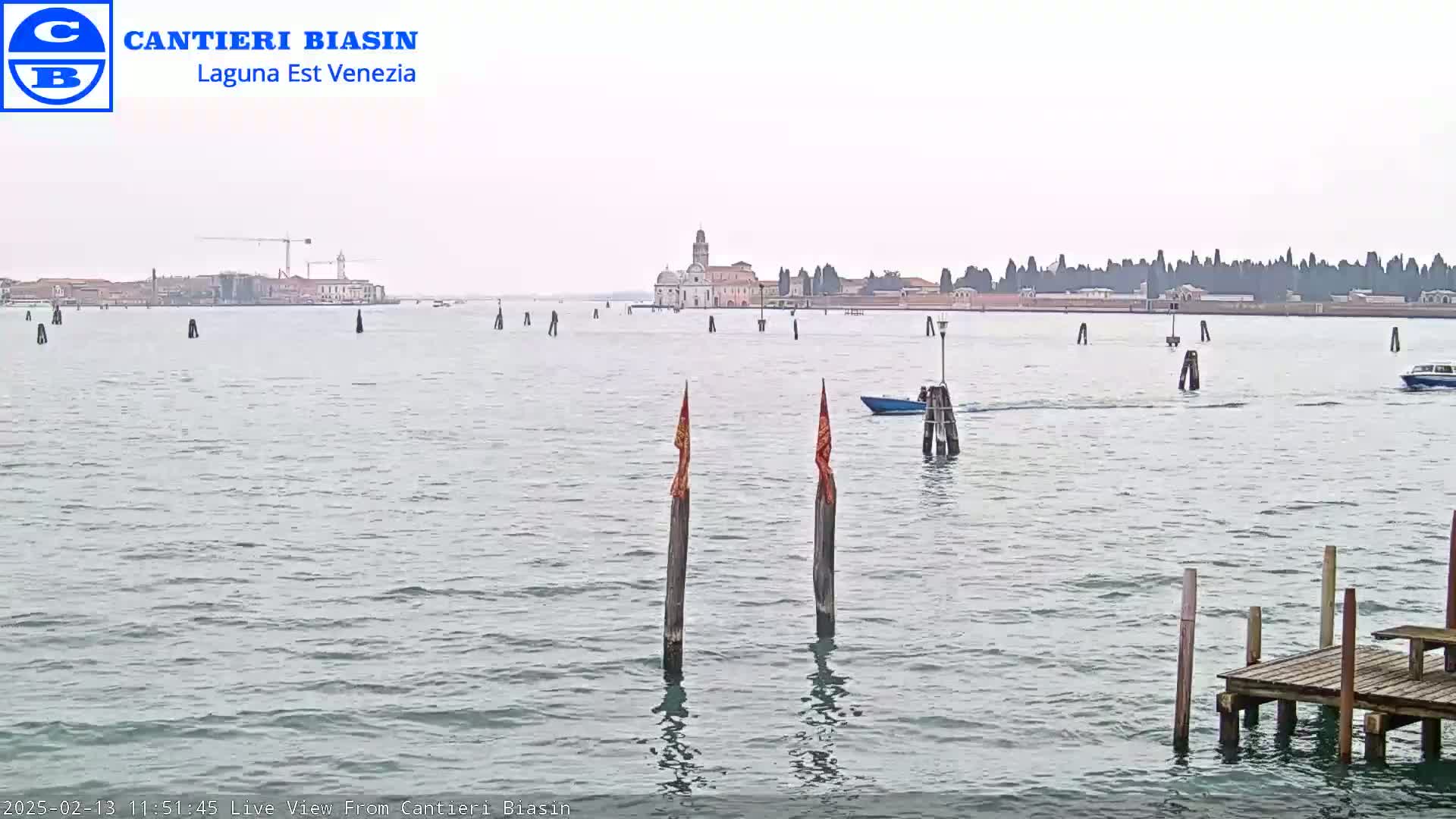 A calm, overcast day shows a body of water with several boats, wooden posts, and a distant island featuring buildings and trees.