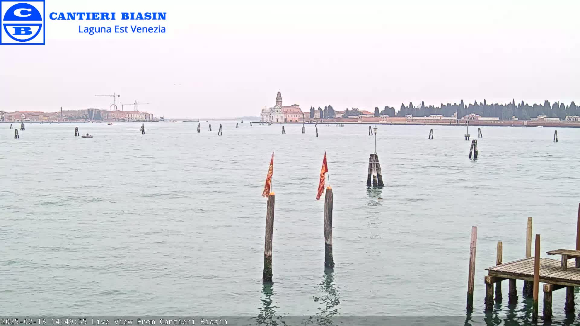 A calm, overcast day shows a body of water with wooden posts and flags, a small dock in the foreground, and a distant island with buildings and trees in the background.