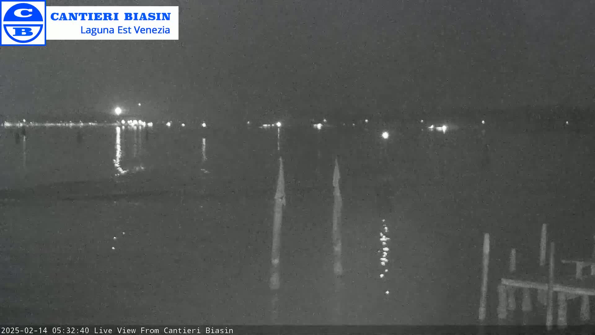A nighttime view of a calm body of water with distant lights reflecting on its surface, and several posts or pilings visible in the foreground.