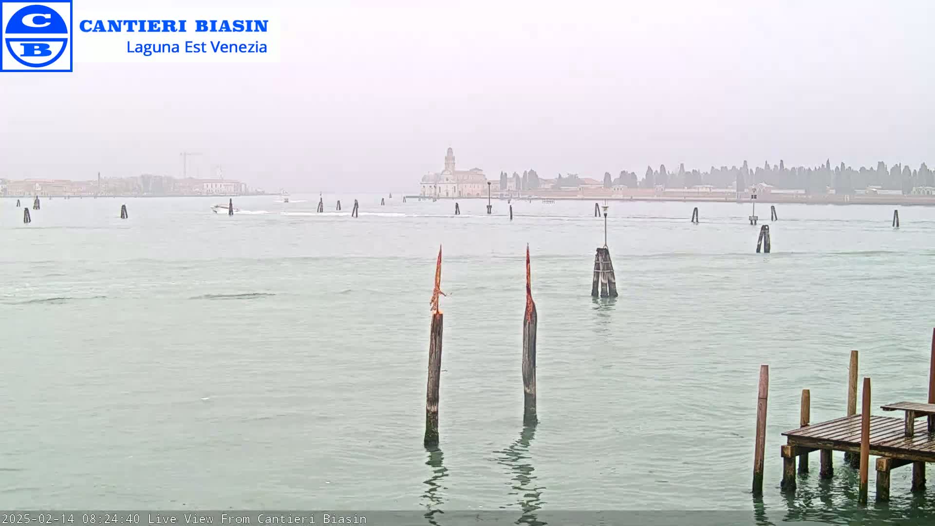 A hazy view of a calm body of water shows a distant cityscape, several wooden posts in the foreground, and a small boat.